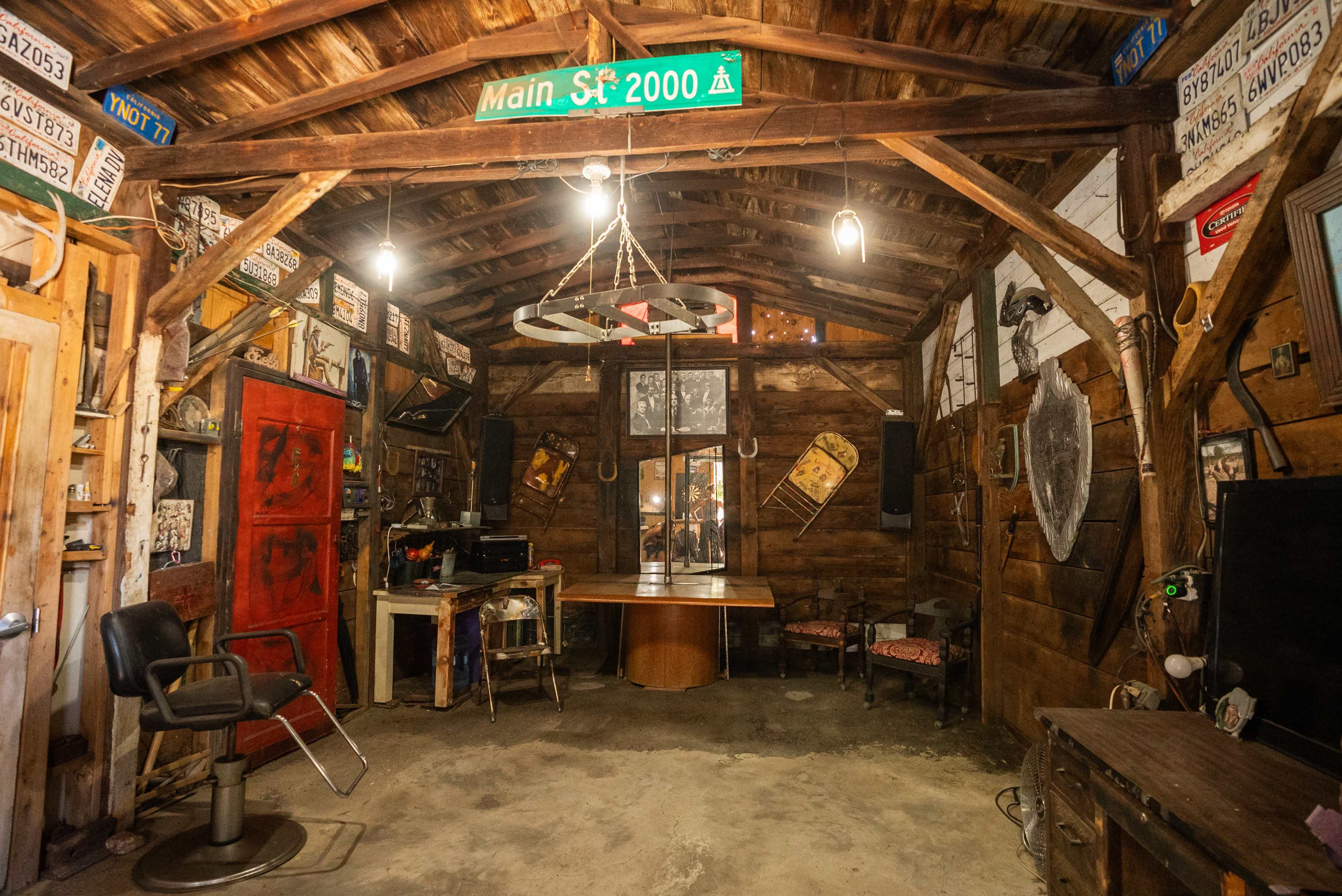 The image shows a rustic, wooden interior of a workshop or garage, featuring various old signs, tools, and furniture, with a prominent red door and a central table.