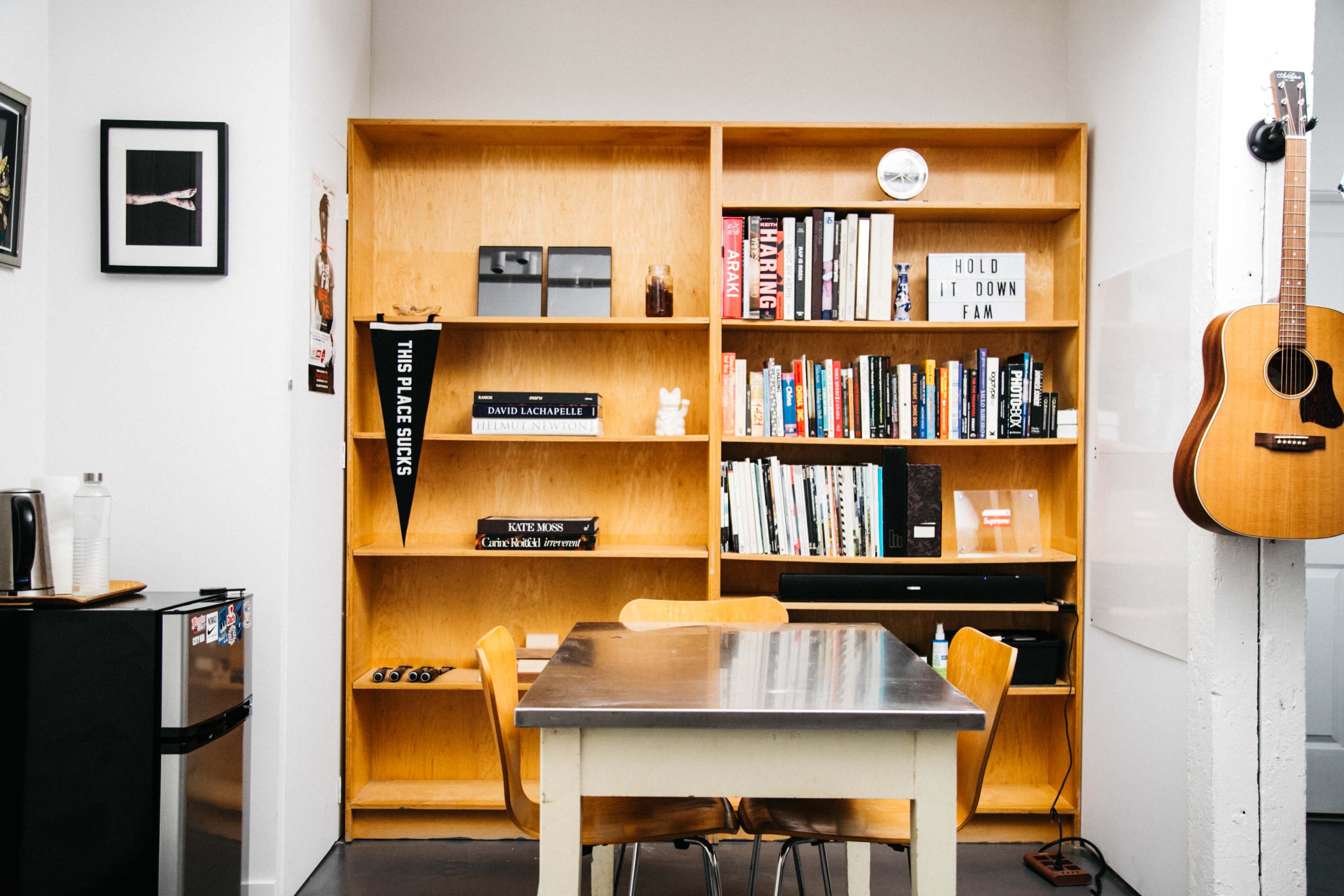 The image depicts a modern kitchen or dining area featuring a wooden bookshelf filled with books, a small table with chairs, and an acoustic guitar hanging on the wall.