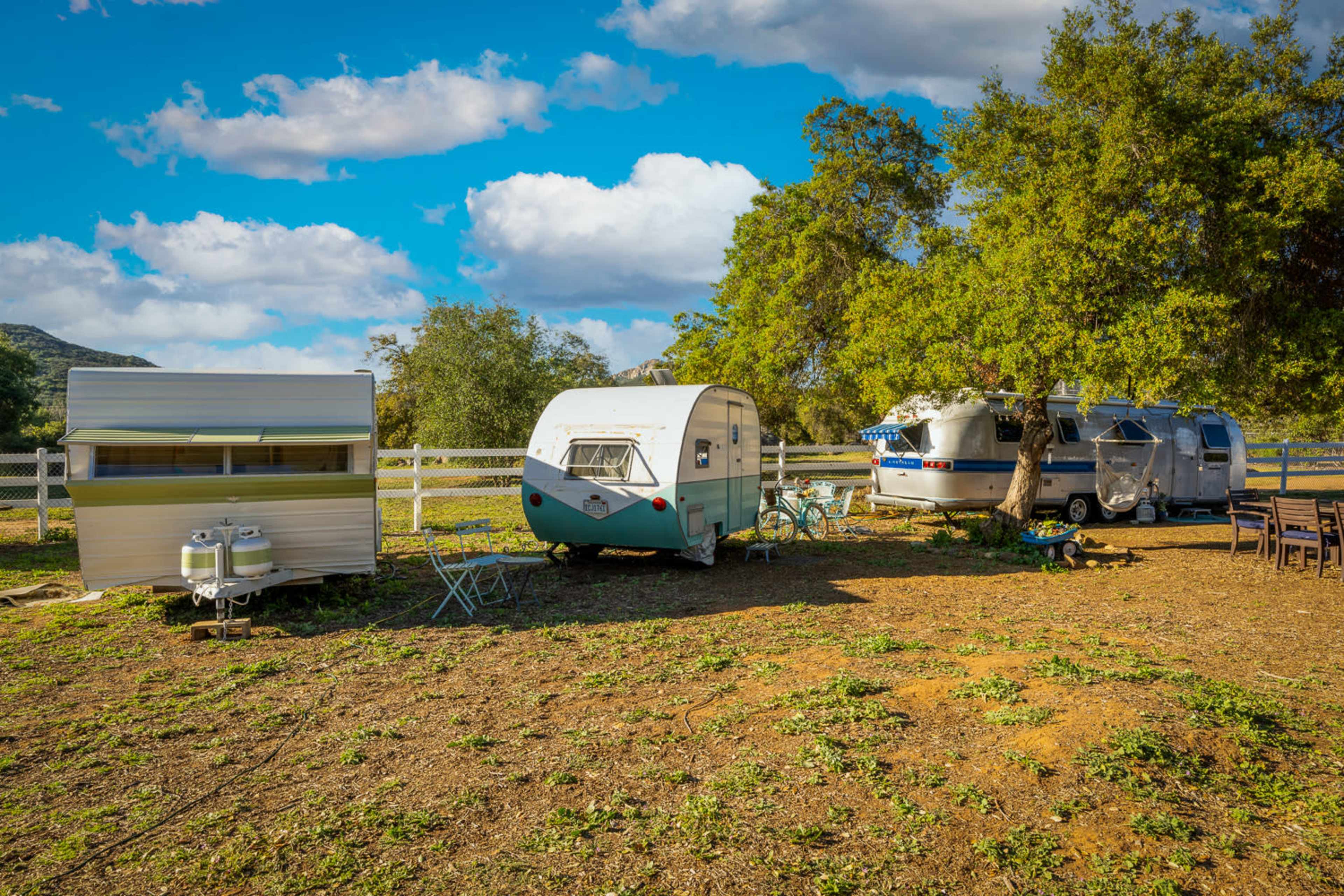Three vintage camper trailers are parked in a grassy area surrounded by trees and a fence under a partly cloudy sky.