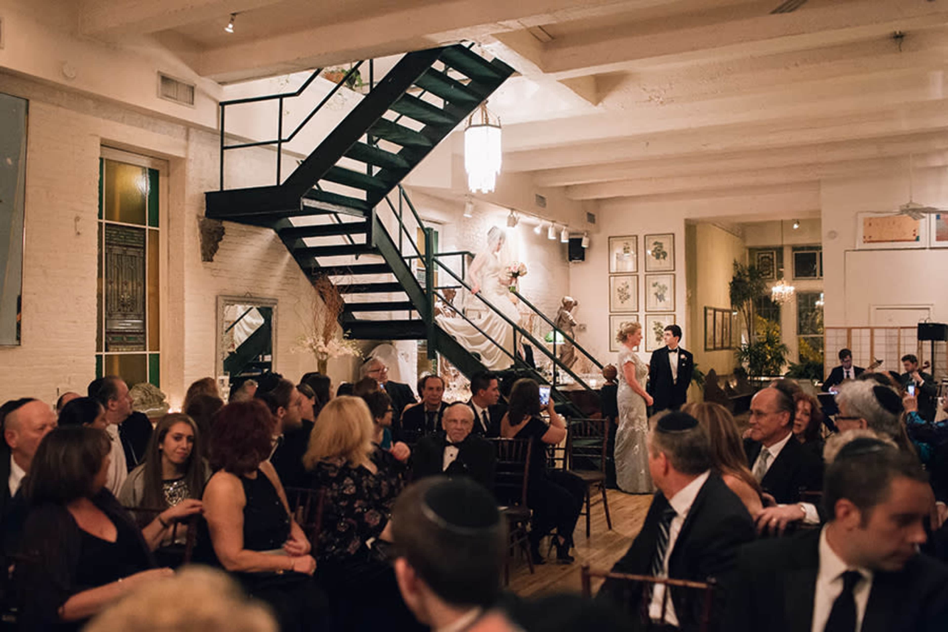 A wedding reception takes place in a spacious venue featuring a staircase and elegantly dressed guests seated at tables.