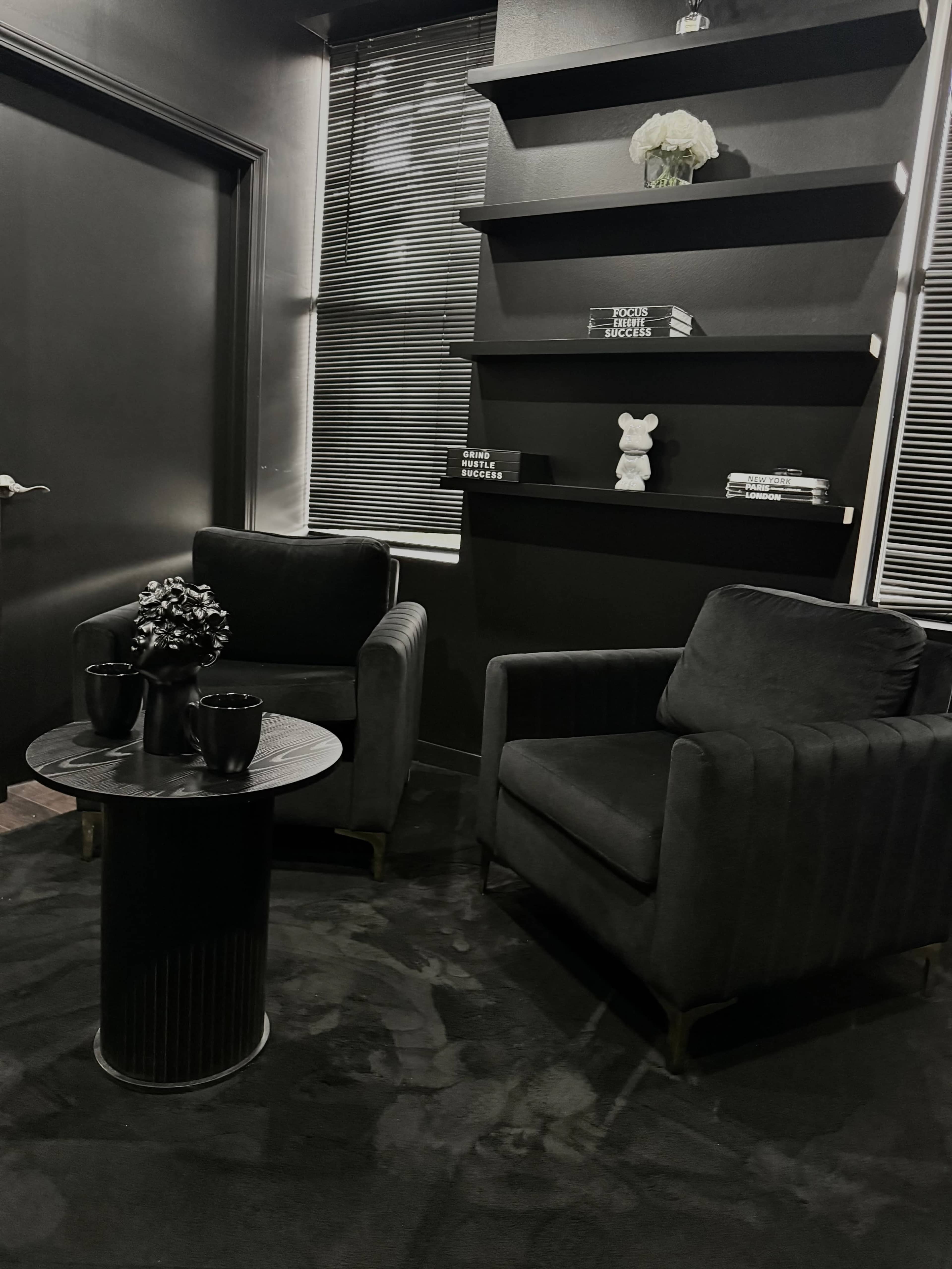 The image shows a small, modern lounge area featuring two black armchairs, a circular black table between them, and a shelf with decorative items against a dark wall.