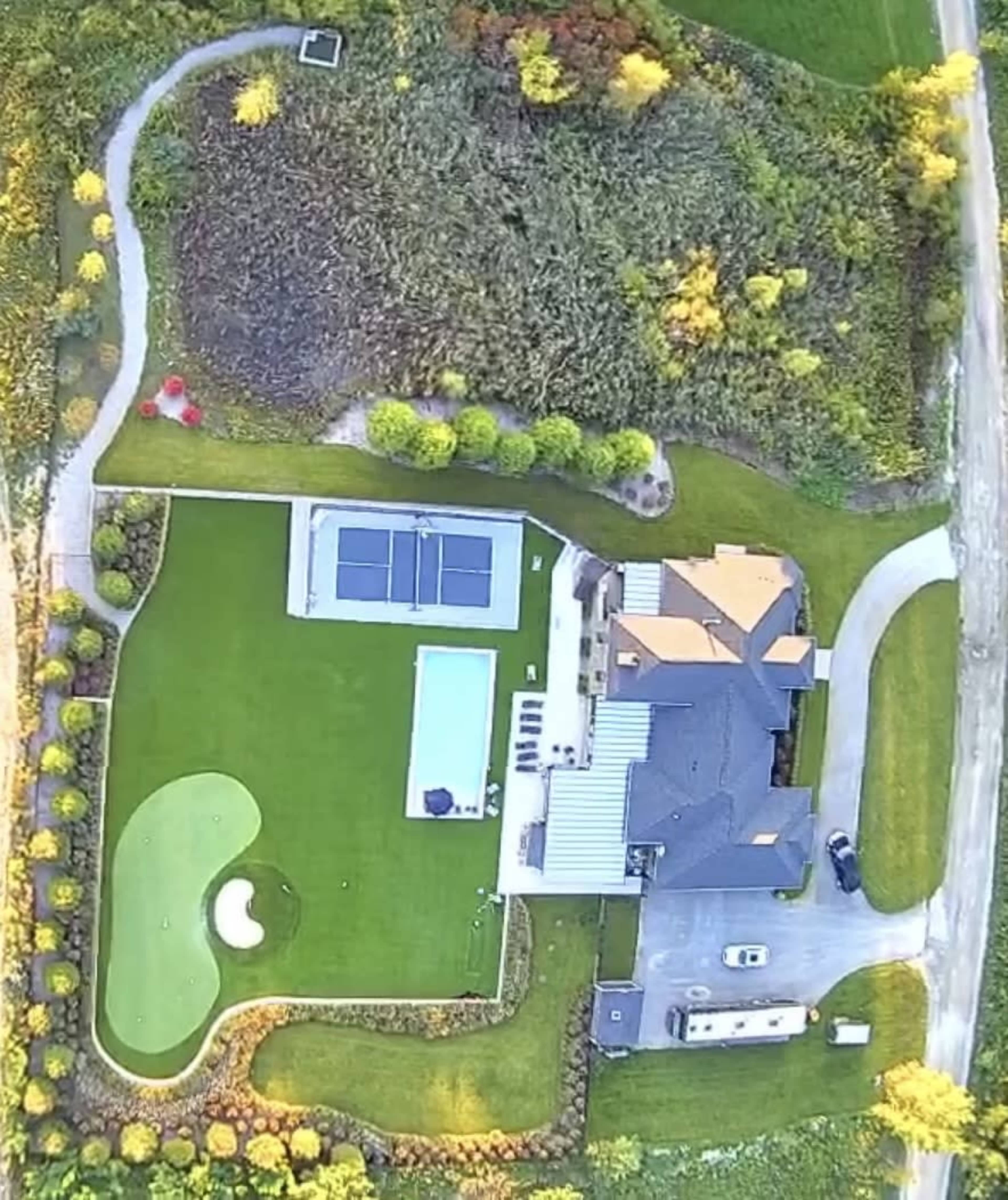 An aerial view of a landscaped property featuring a home, a swimming pool, a tennis court, and a putting green on a large lawn.