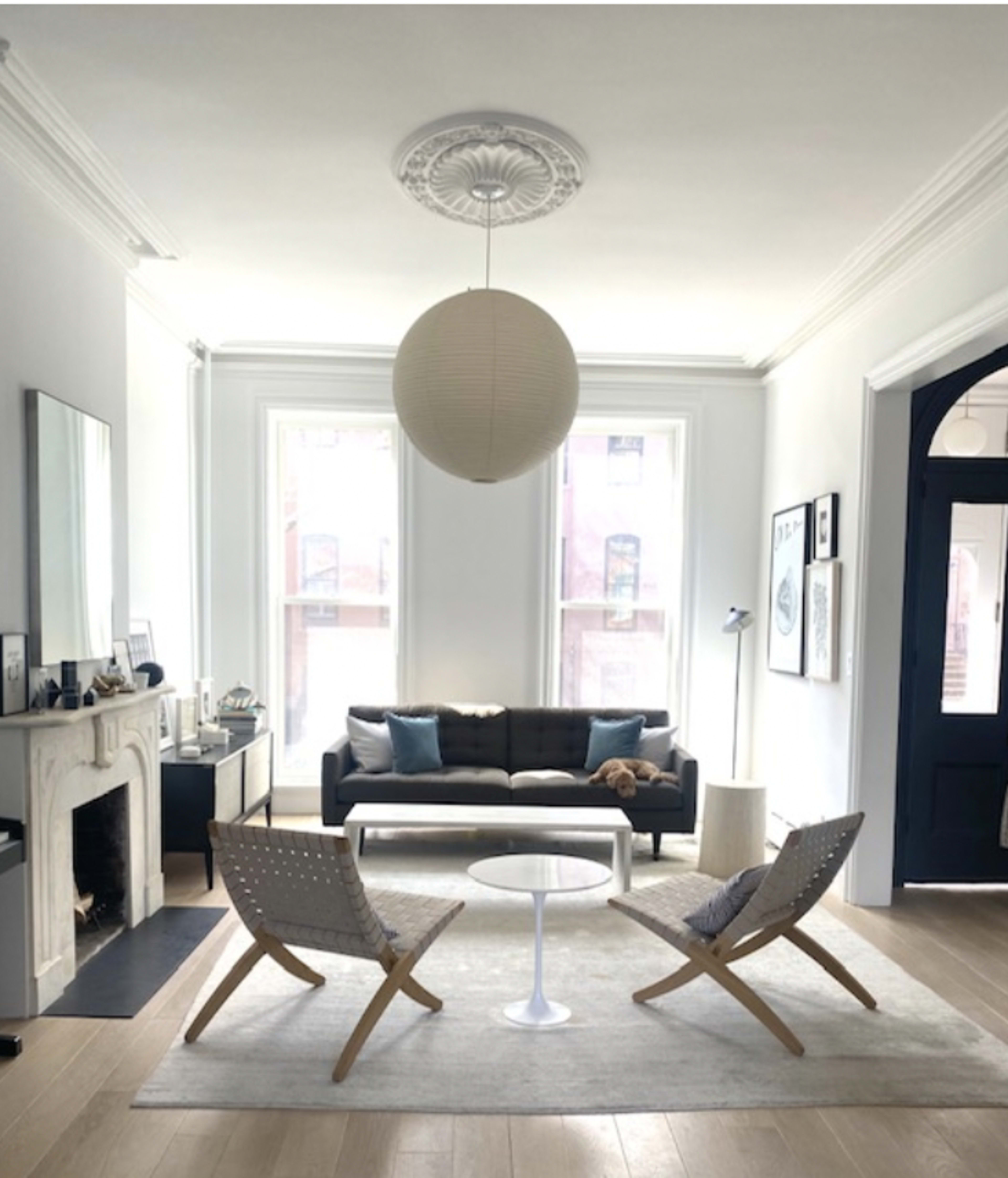 A modern living room with a black sofa, a white circular coffee table, two woven chairs, and large windows allowing natural light.