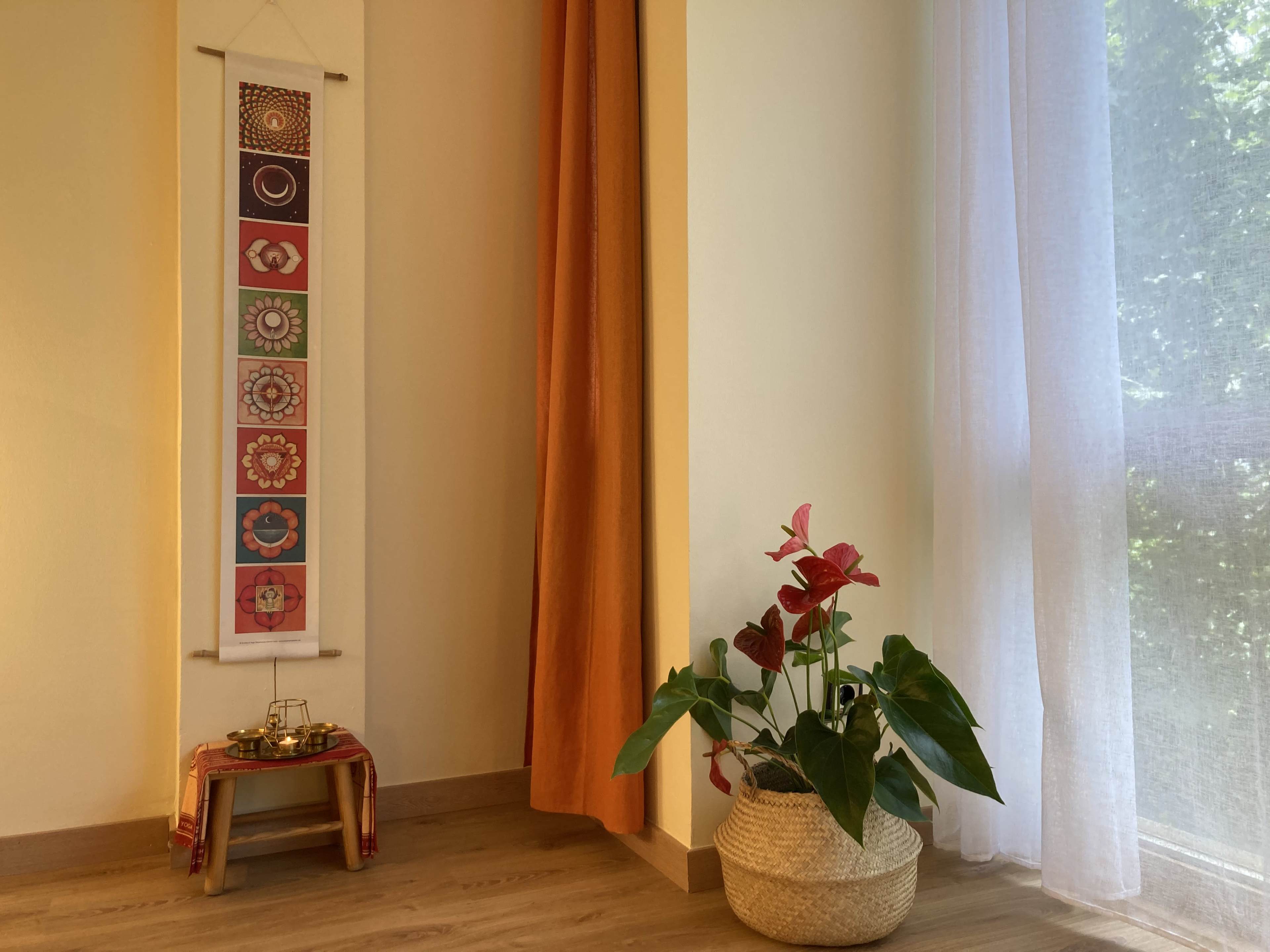 A corner of a room with a wall hanging featuring floral designs, a potted plant, and a small table set with decorative items near a window.