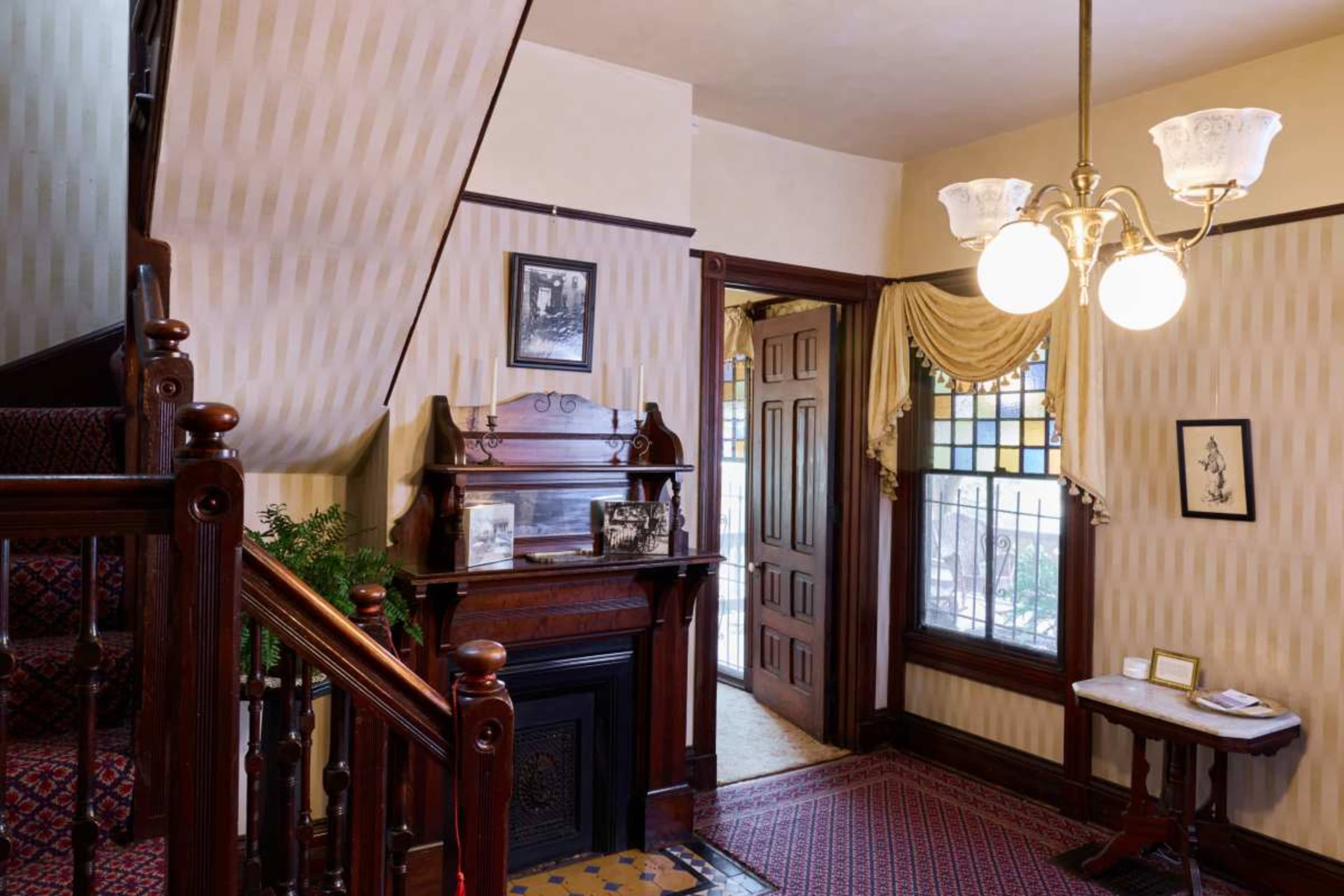 The image shows an elegant entryway with a wooden staircase, a decorative fireplace, and a door leading to another room, all featuring vintage furnishings and patterned wallpaper.