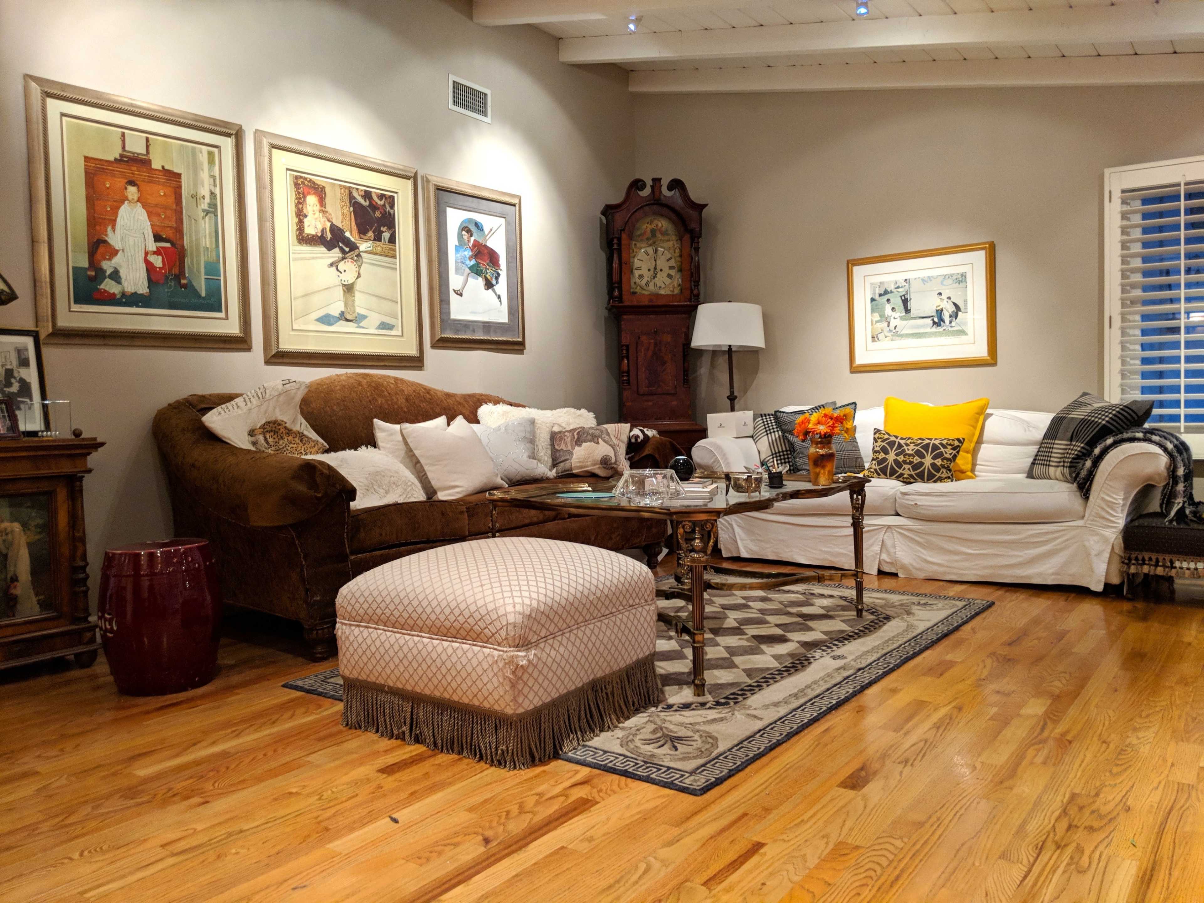 The living room features a mix of furniture including a large brown couch, a white sofa, a wooden coffee table, and an ottoman, all adorned with framed artwork on the walls.