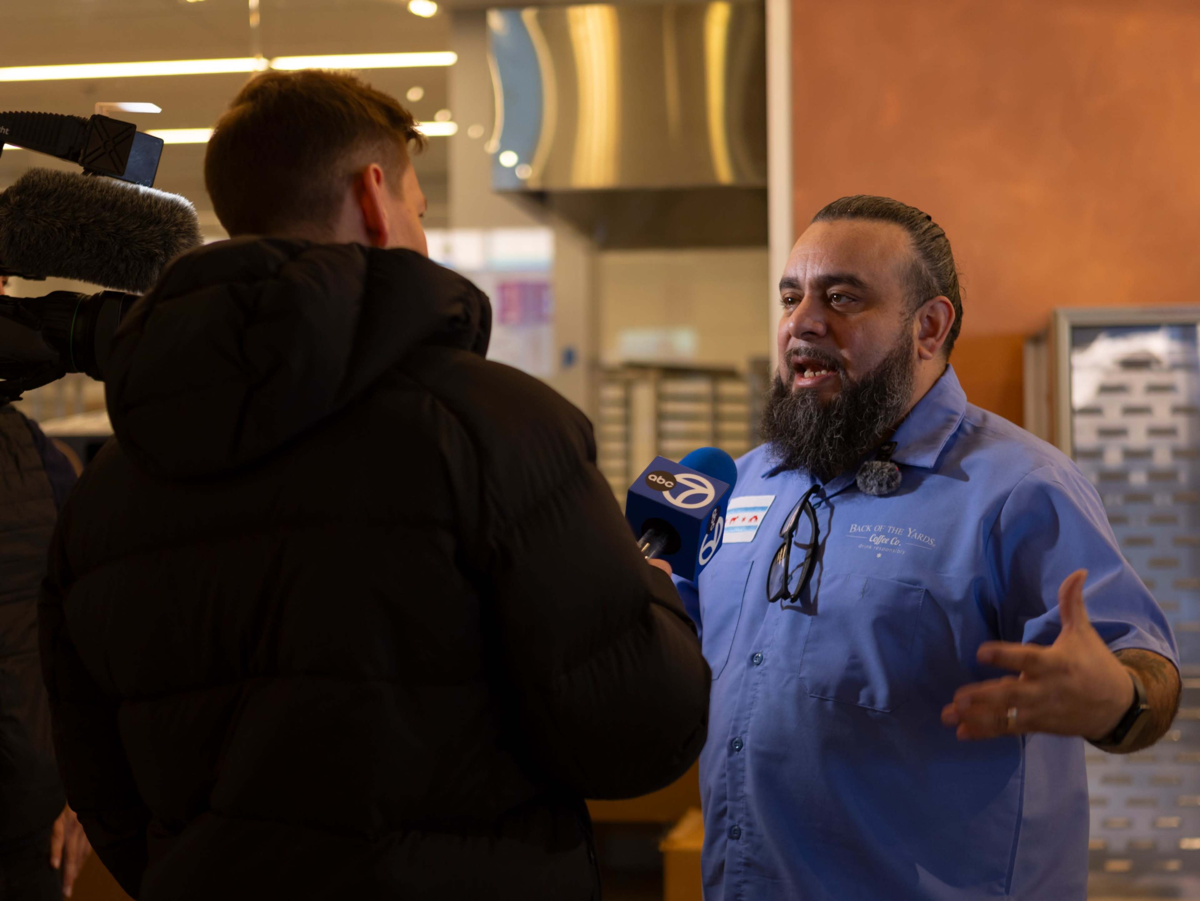 A man with a beard and blue shirt speaks during an interview while a camera operator records nearby.