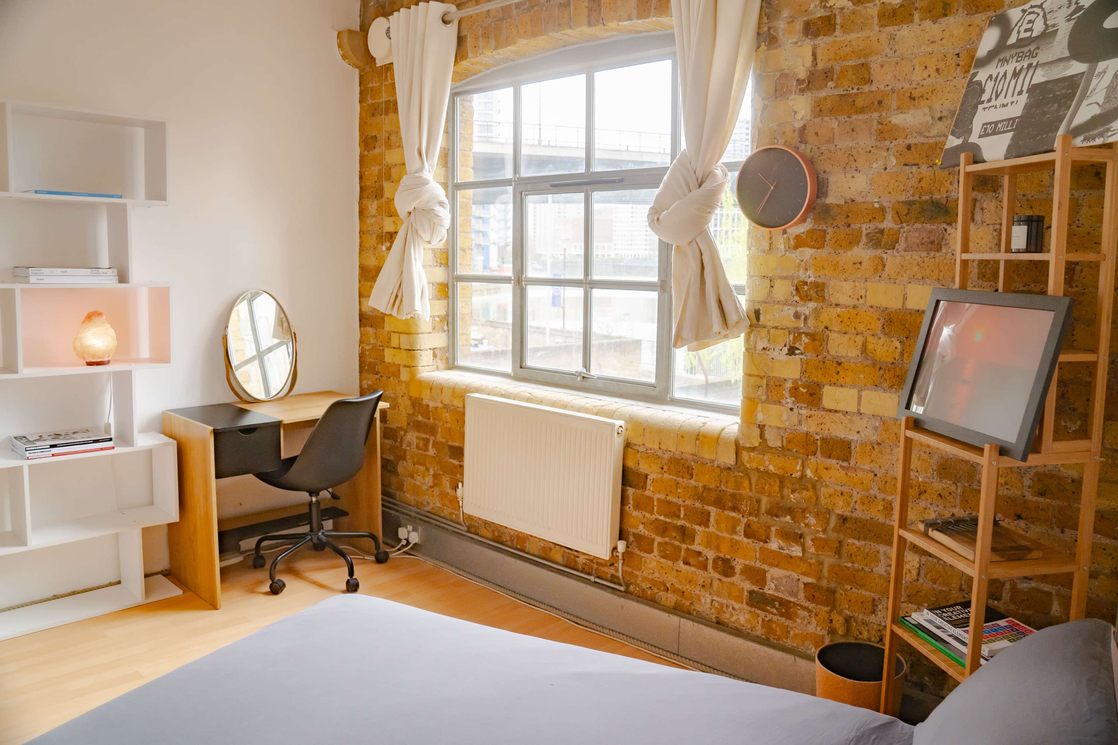 A room features a window with light-colored curtains, a wooden desk with a chair, a bookshelf, and exposed brick walls.