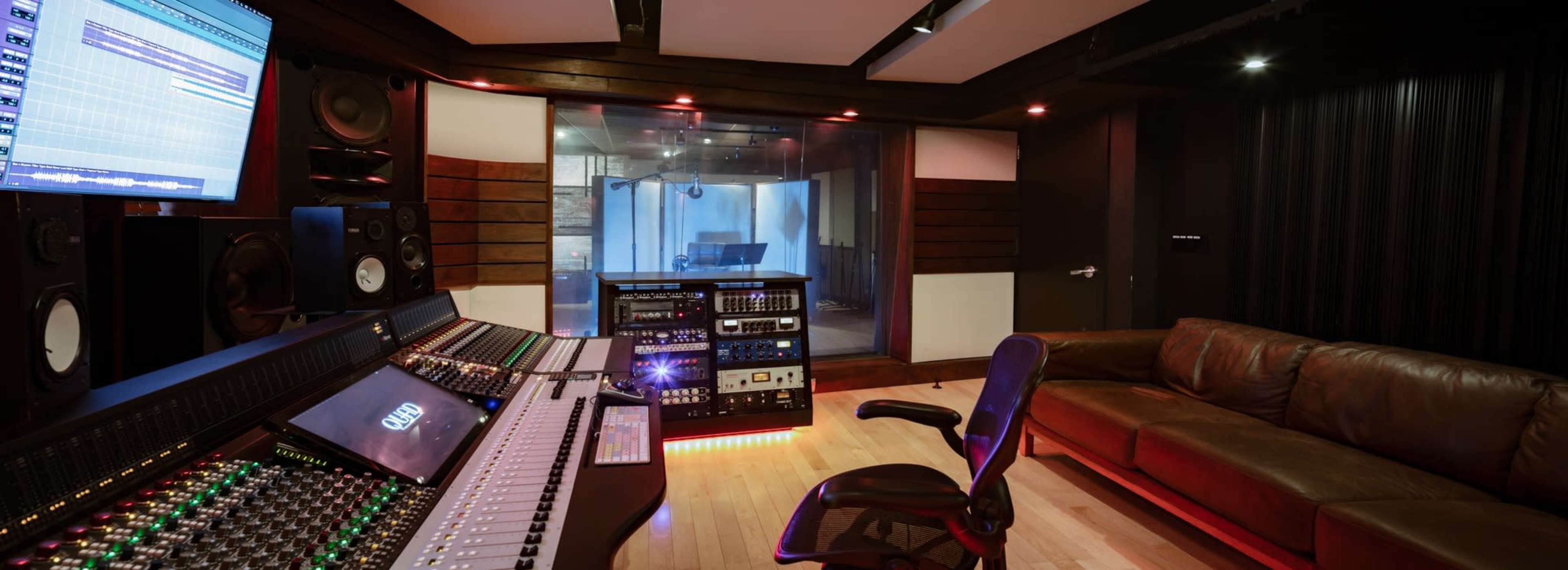 The image shows a modern music studio featuring a mixing console, a computer monitor, and a glass-walled recording booth.