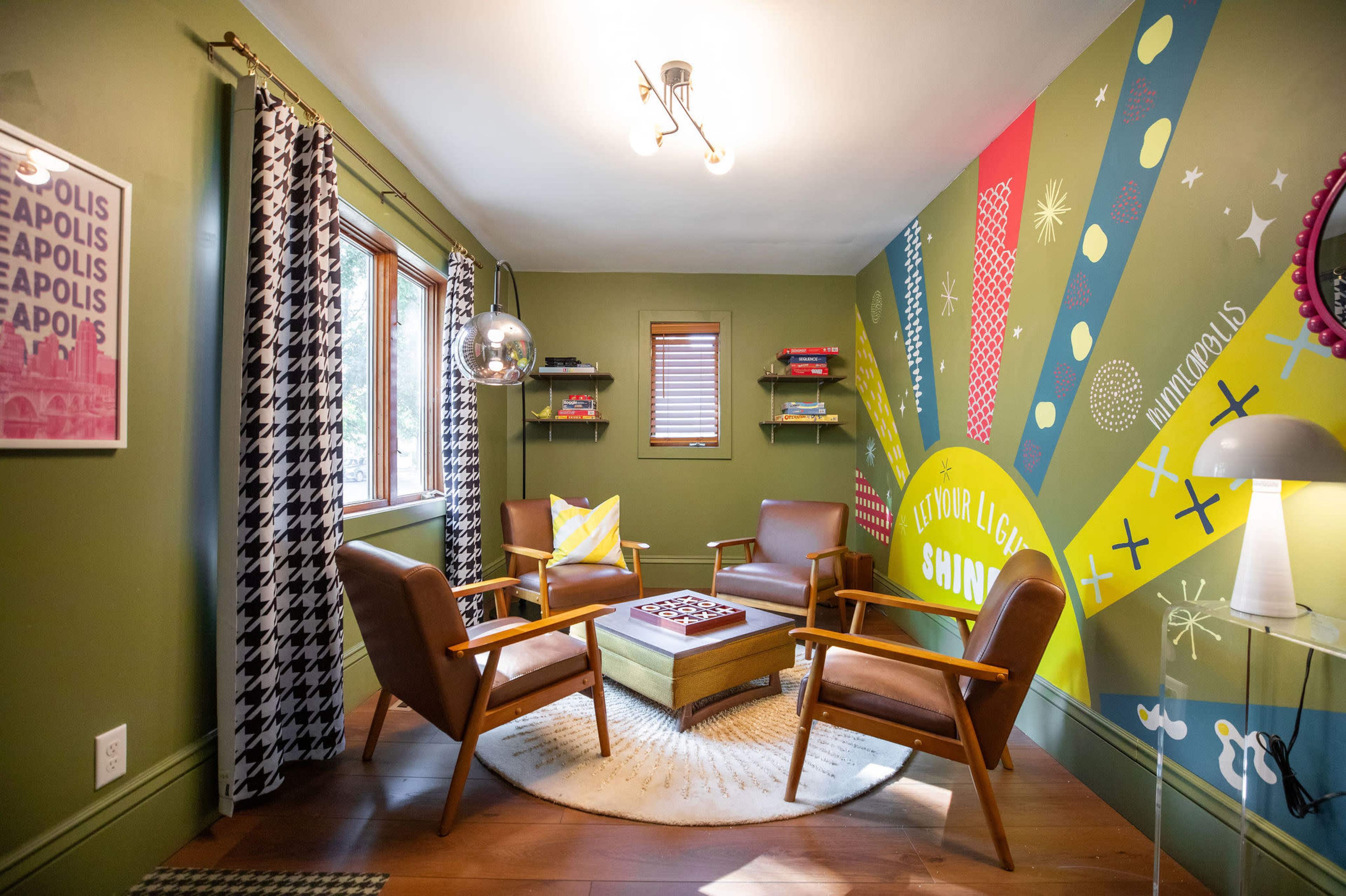 A small room with green walls, two wooden chairs, a round table with a game board, and decorative elements, including a bold mural and patterned curtains.