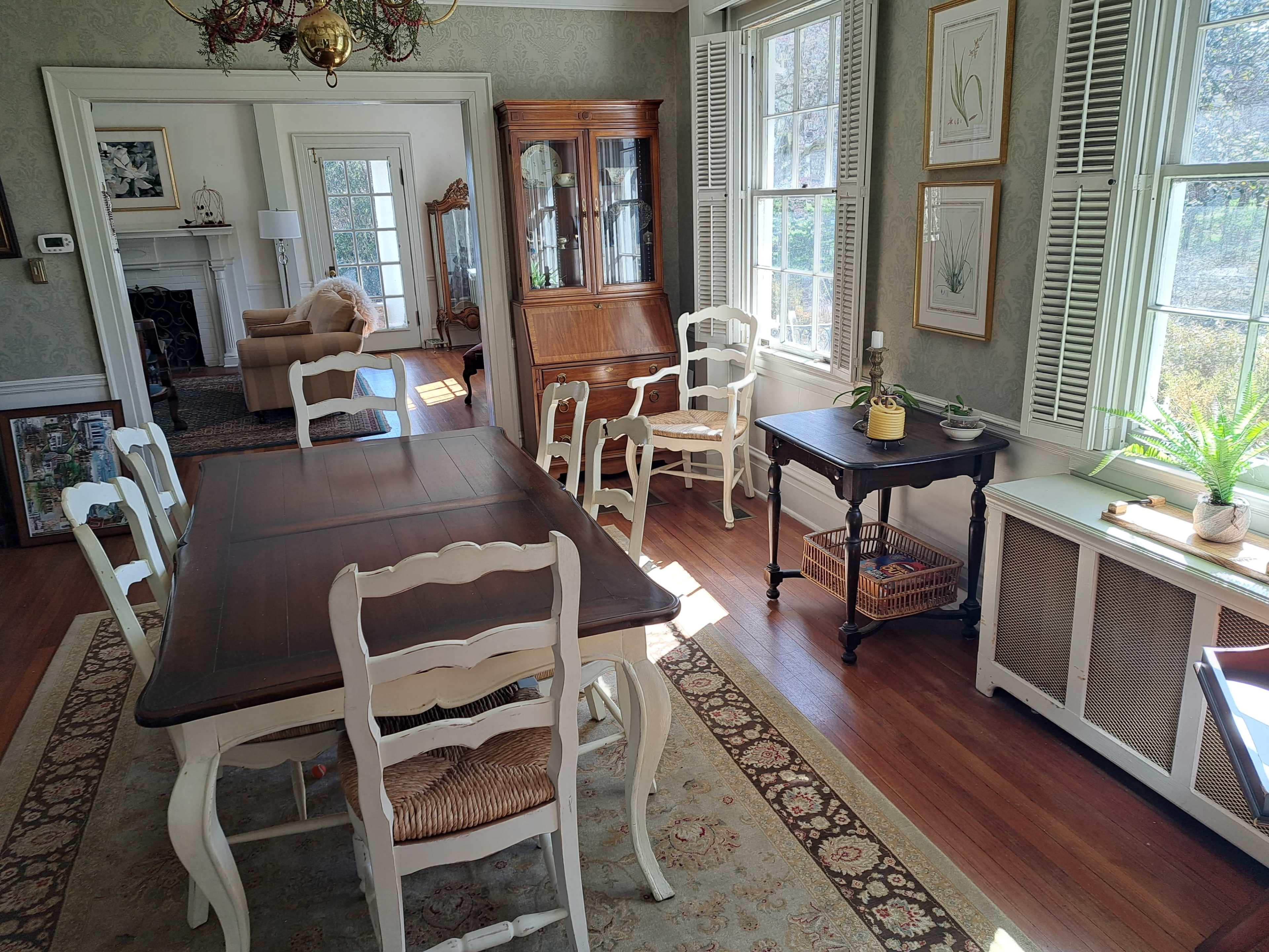 The image shows a sunlit dining room with a wooden table surrounded by white chairs, a sideboard, and large windows featuring decorative plants.