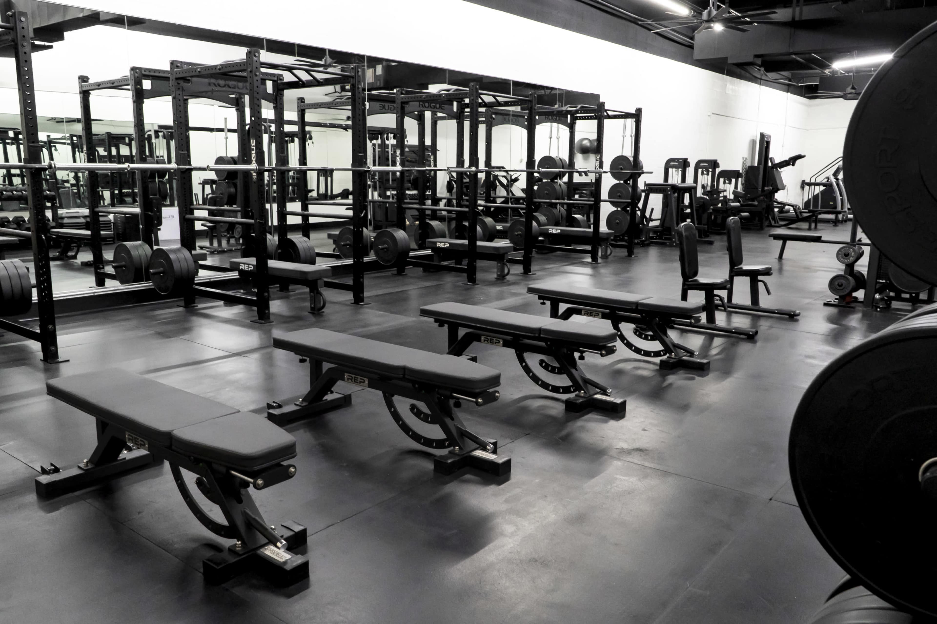 The image shows a modern gym interior with multiple weight benches and various workout stations arranged in a spacious area.