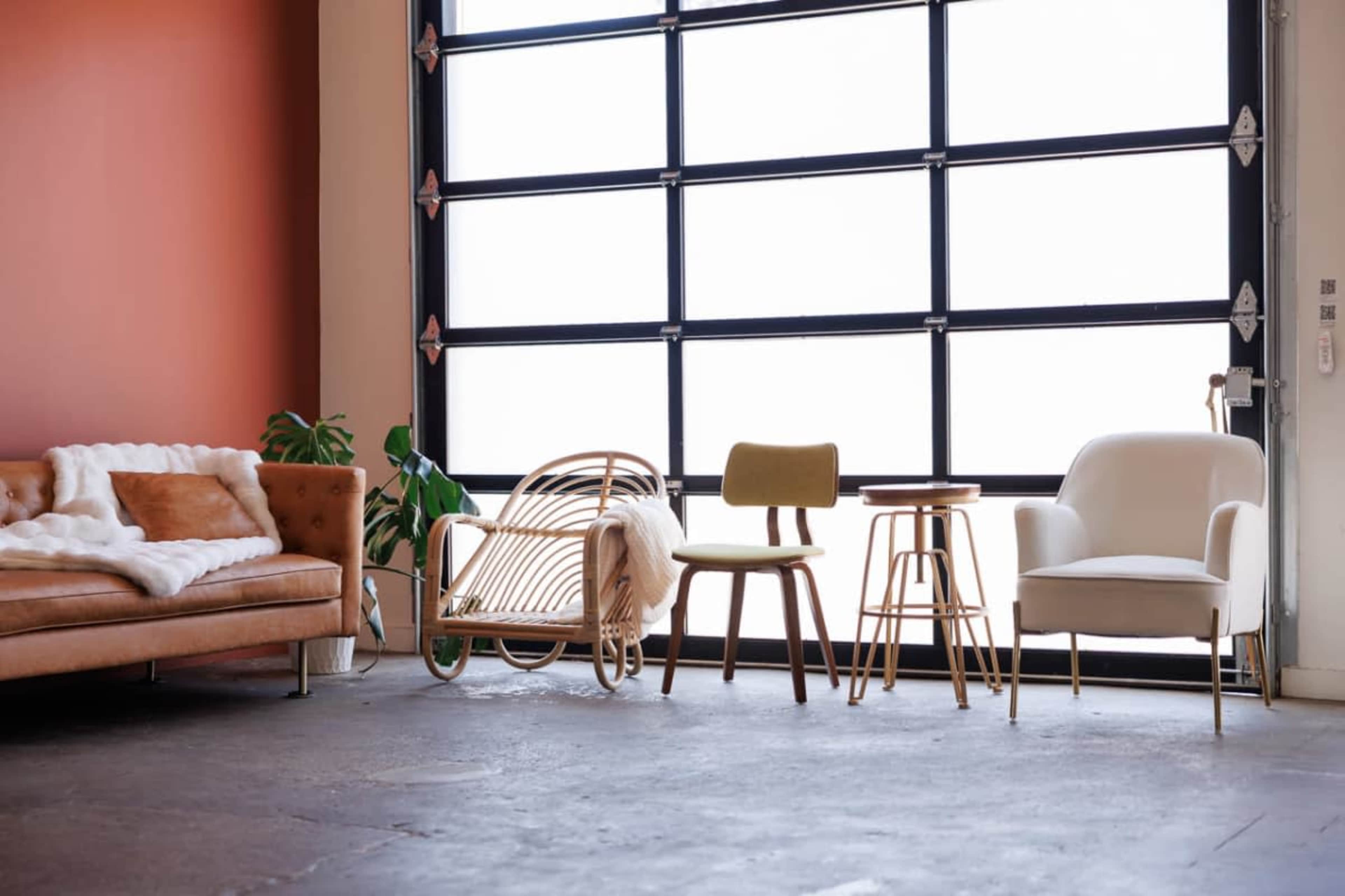 The image shows a minimalistic interior space featuring a brown leather sofa, a rattan chair, a green upholstered chair, a small wooden table, and a white armchair, all arranged near a large garage door.