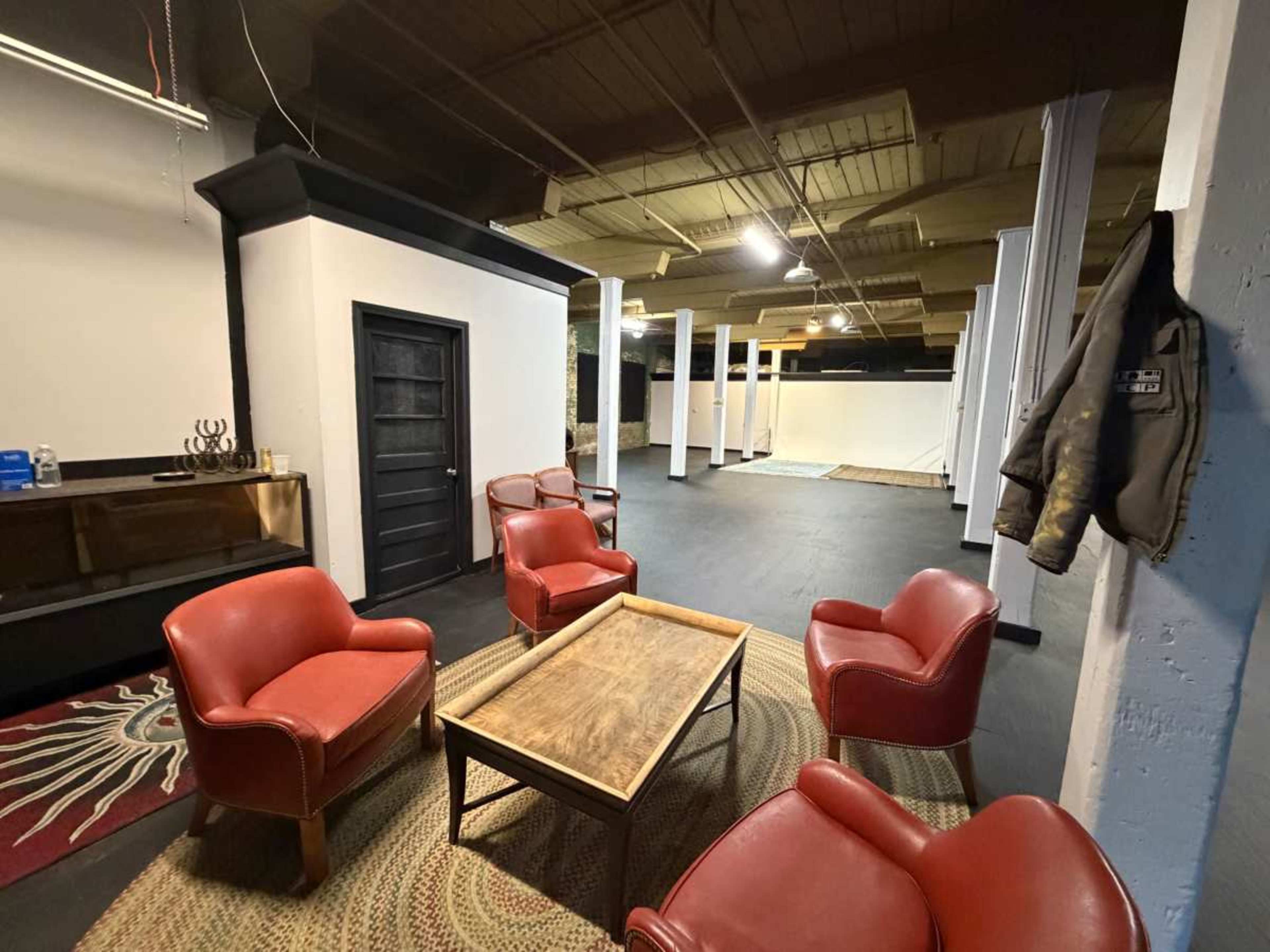 The image shows a spacious room with a few red armchairs surrounding a wooden coffee table, a light-colored rug on the floor, and exposed rafters above.