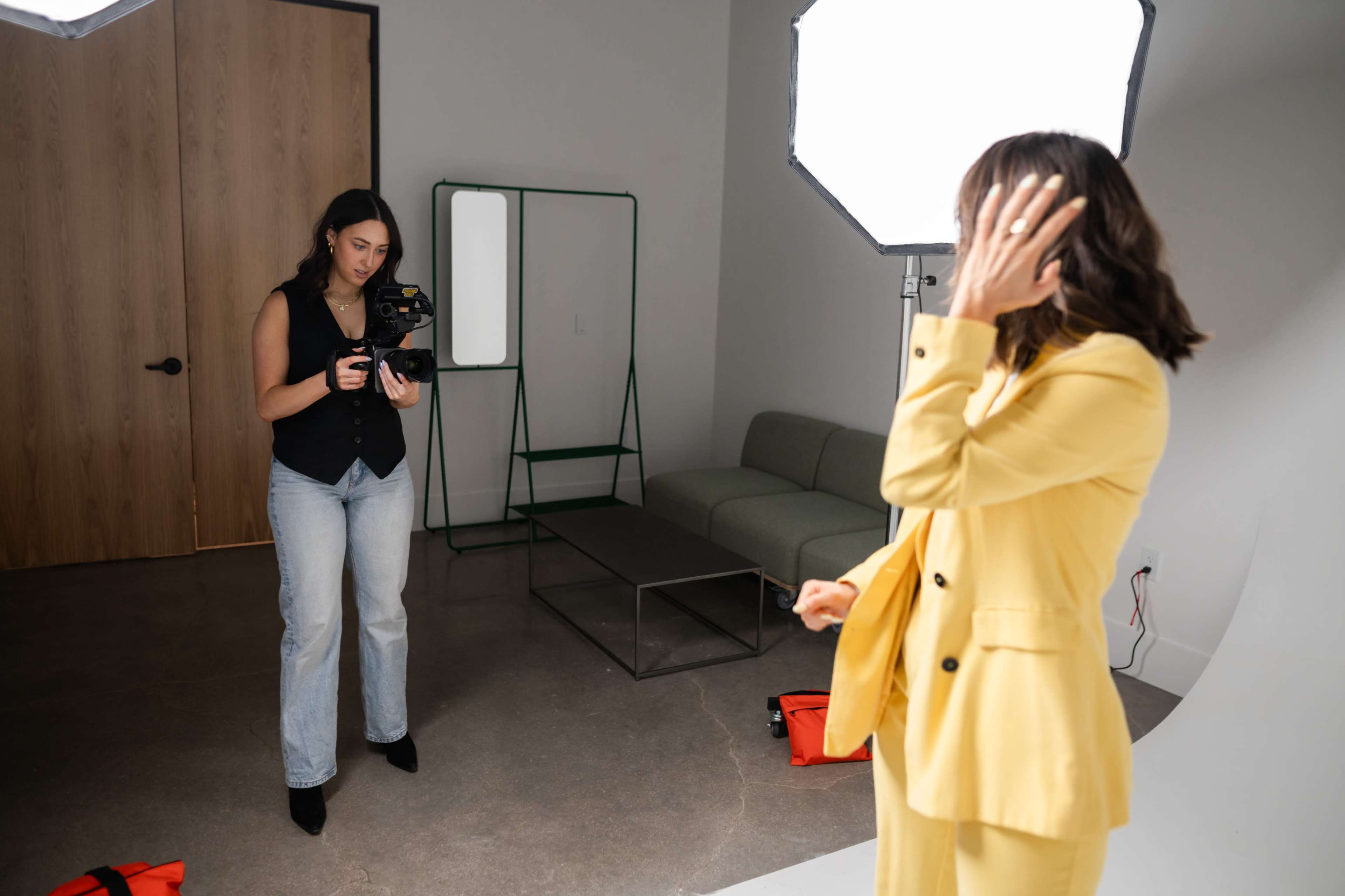 A woman in a yellow suit poses in front of a photographer holding a camera in a well-lit studio.