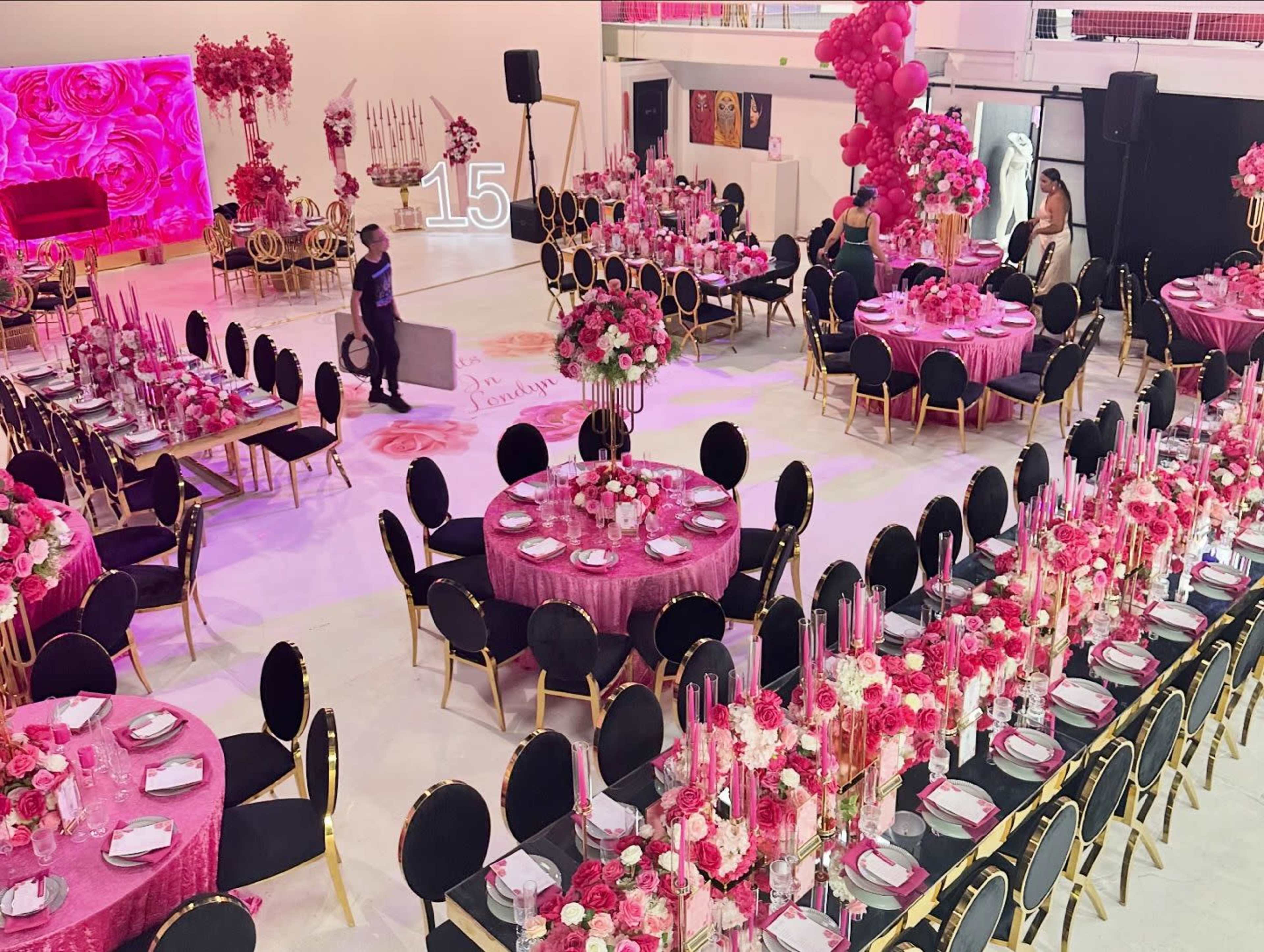 A large event space is set with multiple round and long tables decorated in shades of pink, each adorned with floral arrangements and candles.
