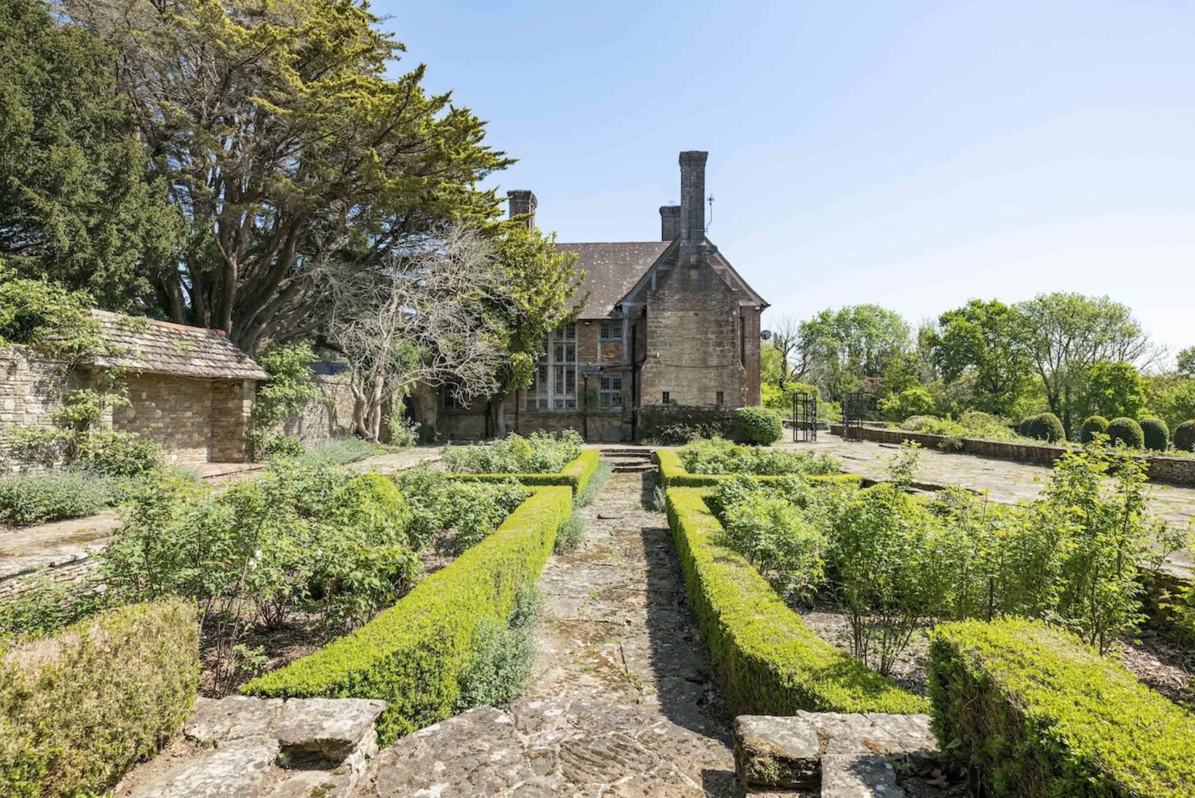 A large stone house stands in the background, surrounded by neatly arranged green hedges and a garden path in the foreground.