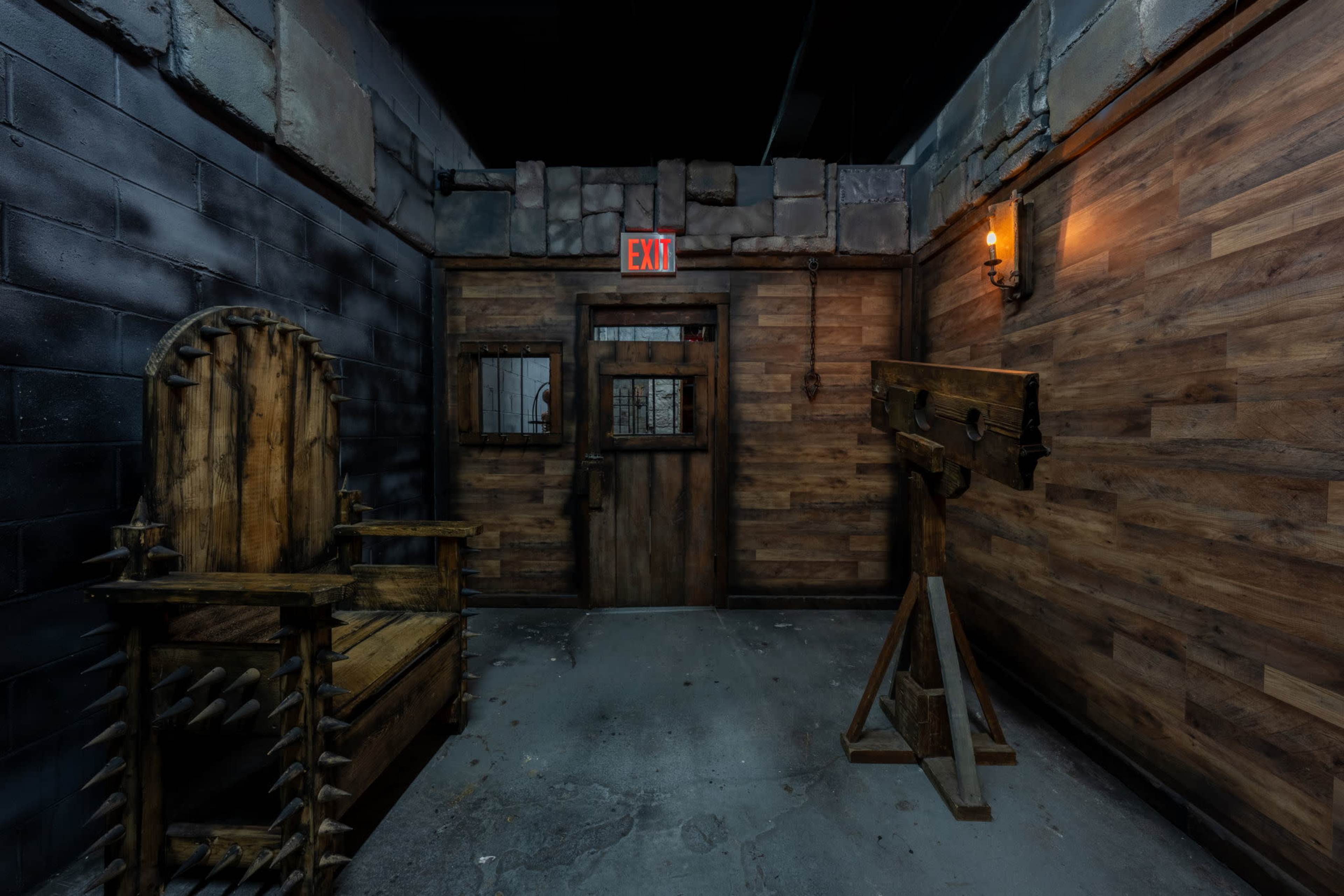 The image shows a dimly lit room with a wooden chair featuring spikes, a stocks device, and a wooden door with an exit sign above it.