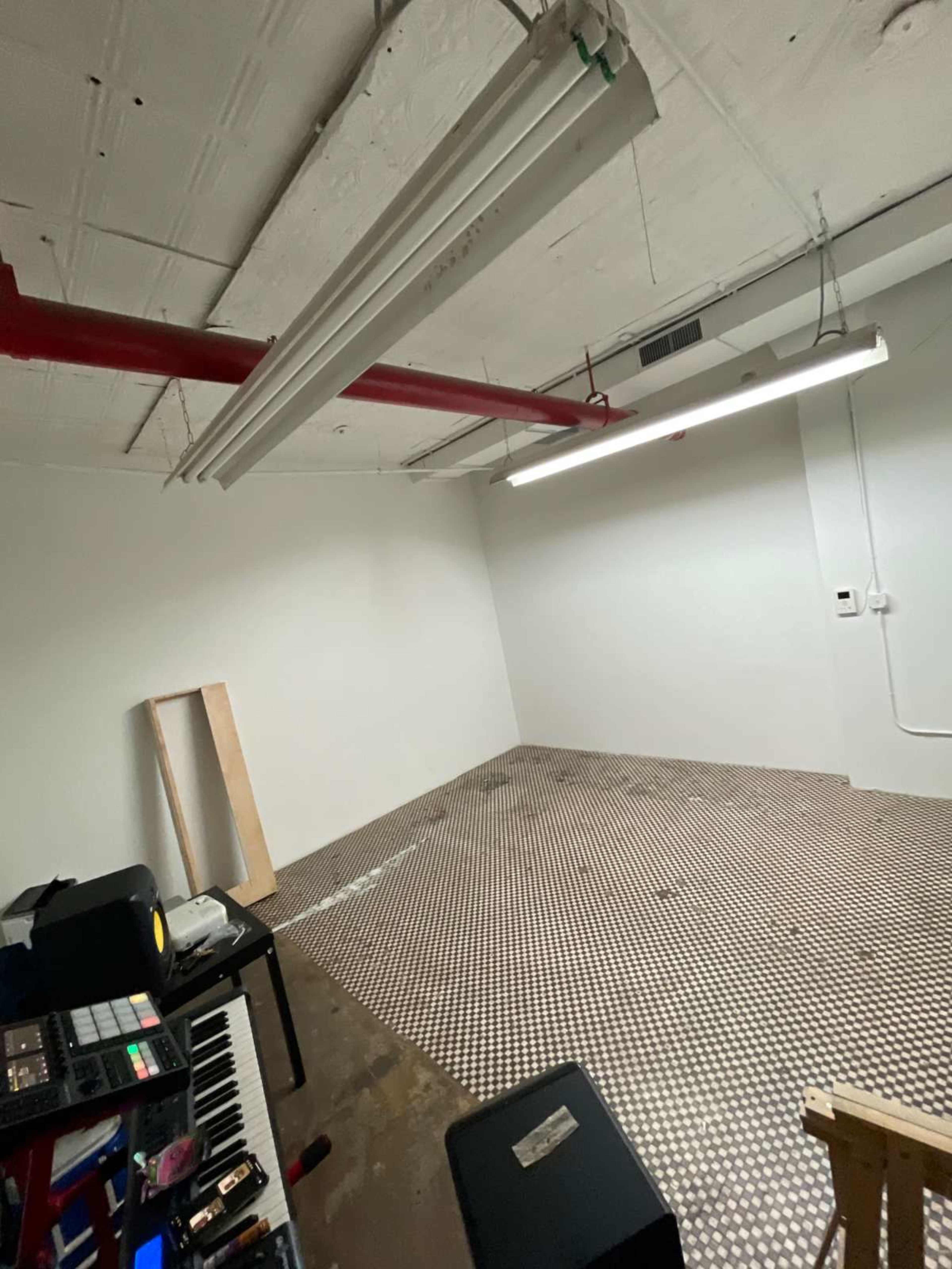 The image shows a sparsely furnished room with a textured floor, exposed ceiling pipes, and minimal equipment on one side.