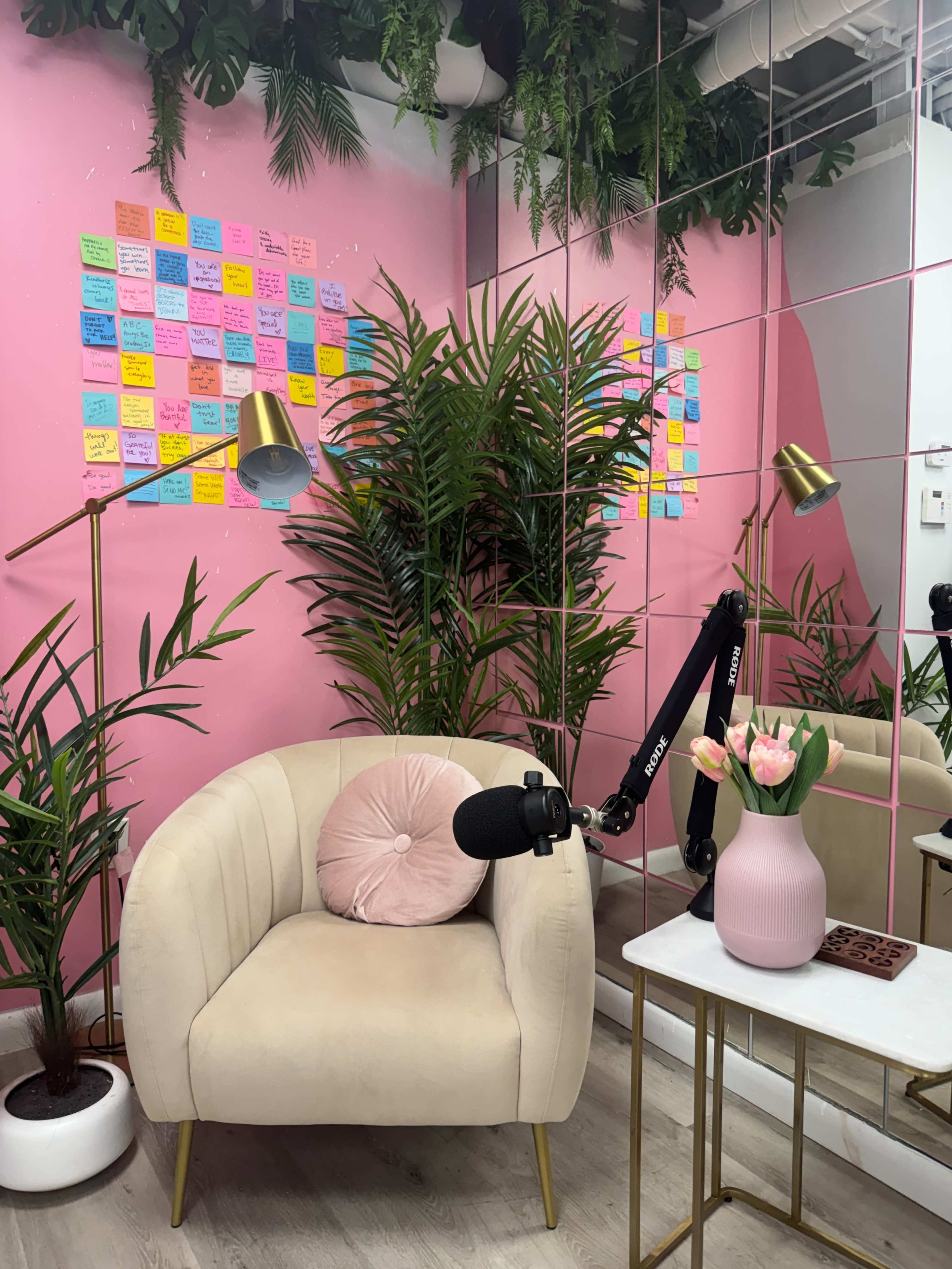 The image features a cozy corner with a pink wall covered in colorful sticky notes, a light-colored armchair, a microphone on an arm stand, a white table with a pink vase of tulips, and potted plants.
