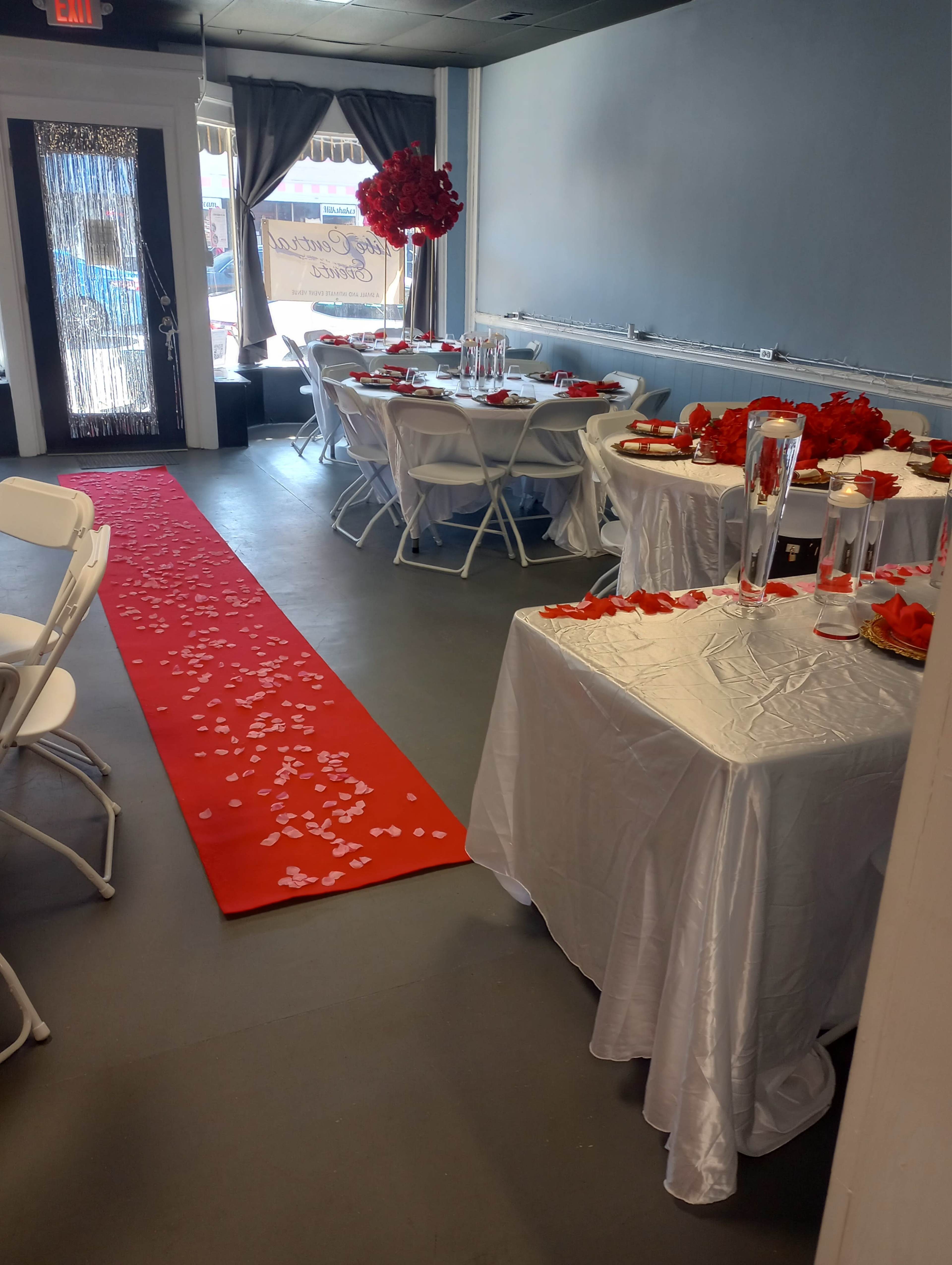 The image shows a decorated interior space set up for an event, featuring white tables with tablecloths, a red carpet with rose petals, and floral arrangements.