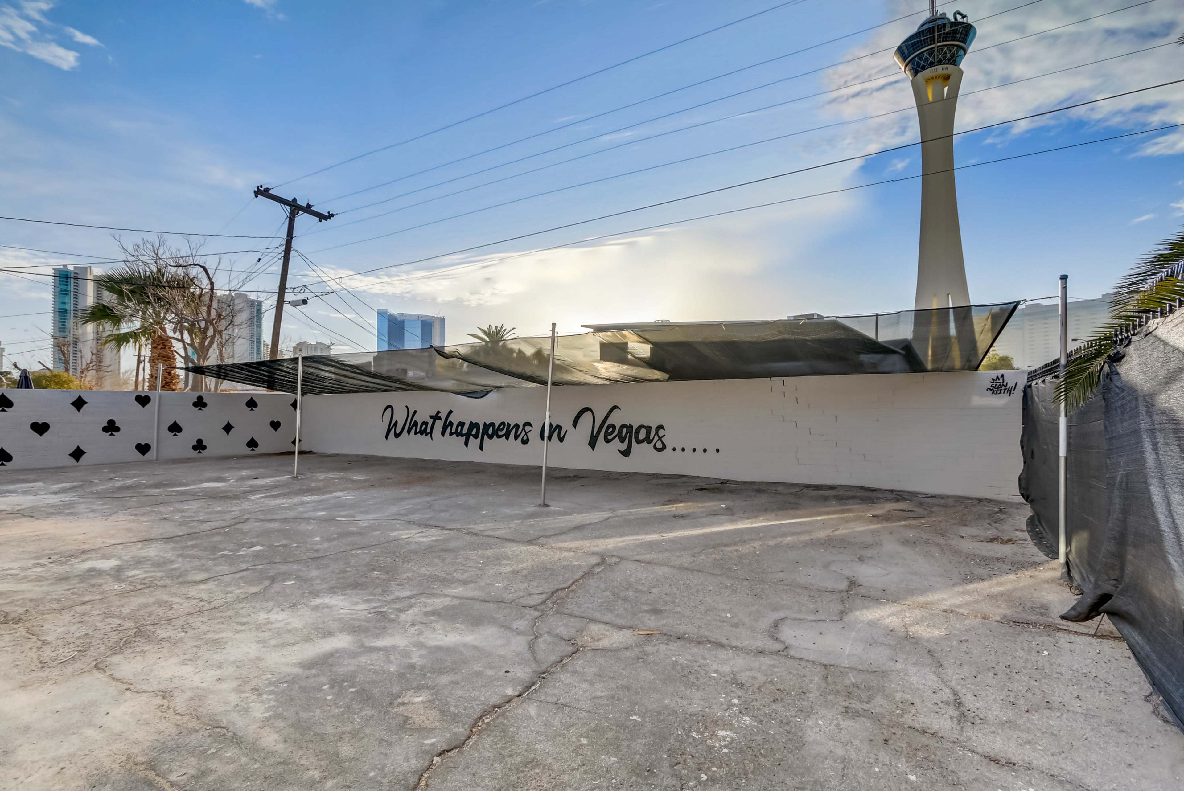 The image shows an empty lot with a mural reading "What happens in Vegas..." on a wall, with the Stratosphere Tower visible in the background.