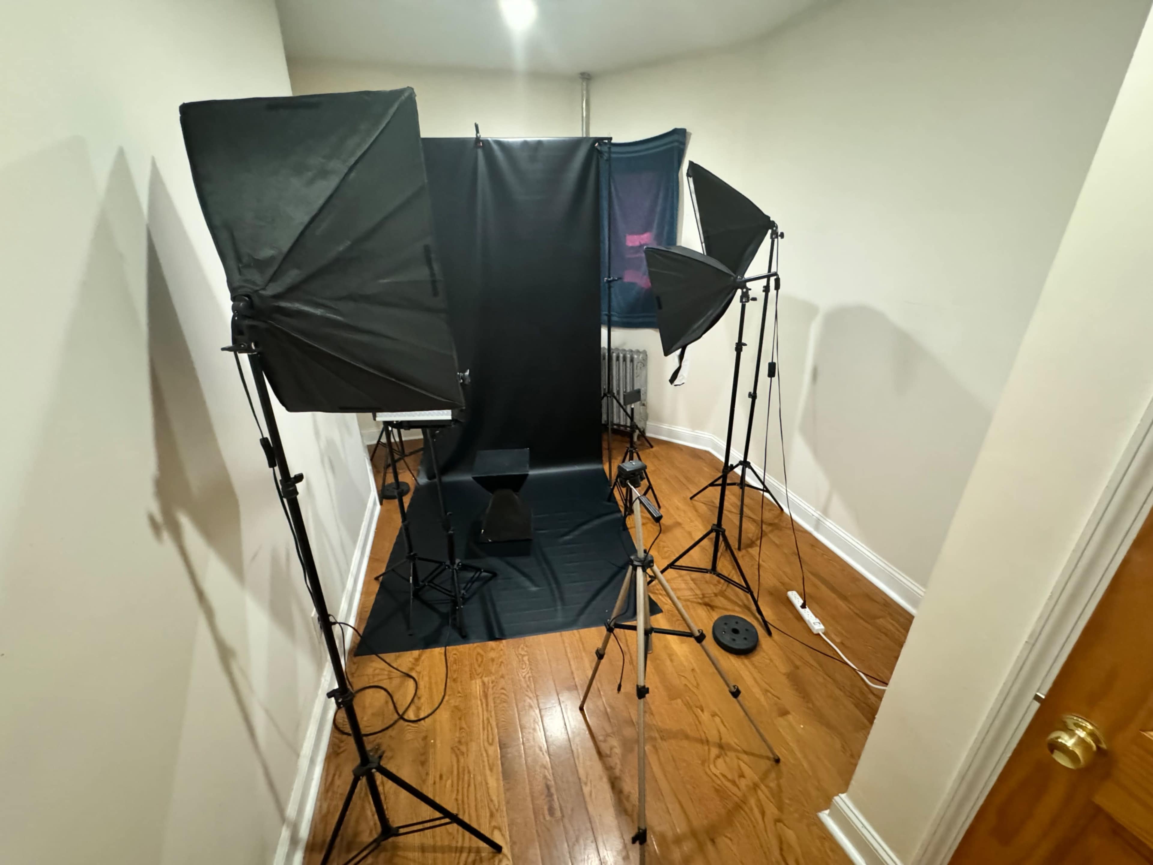 The image shows a photo studio setup with three softboxes, a black backdrop, and a stool on a wooden floor.