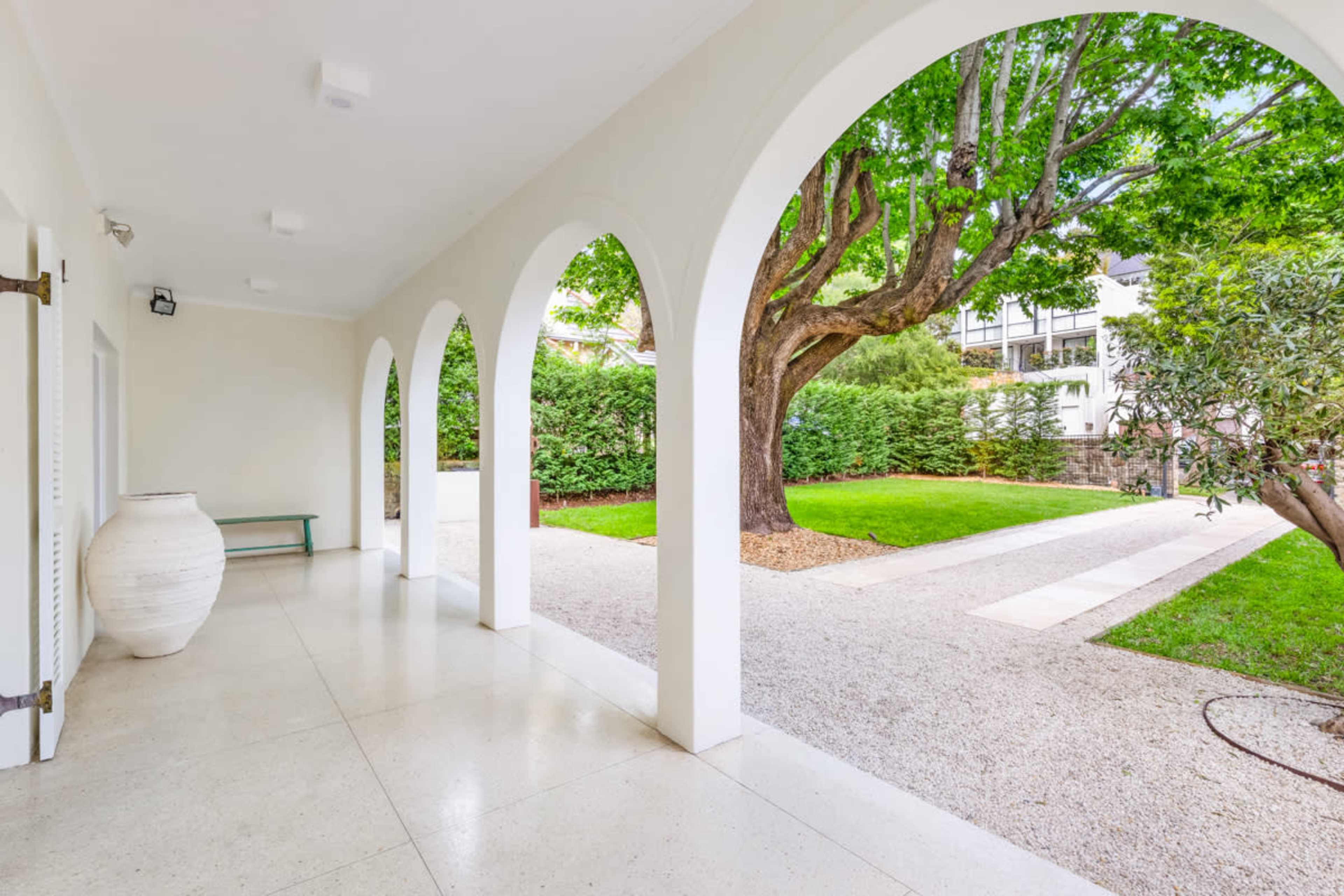 The image depicts a spacious corridor featuring several arches, leading to a landscaped garden with a large tree and decorative elements.