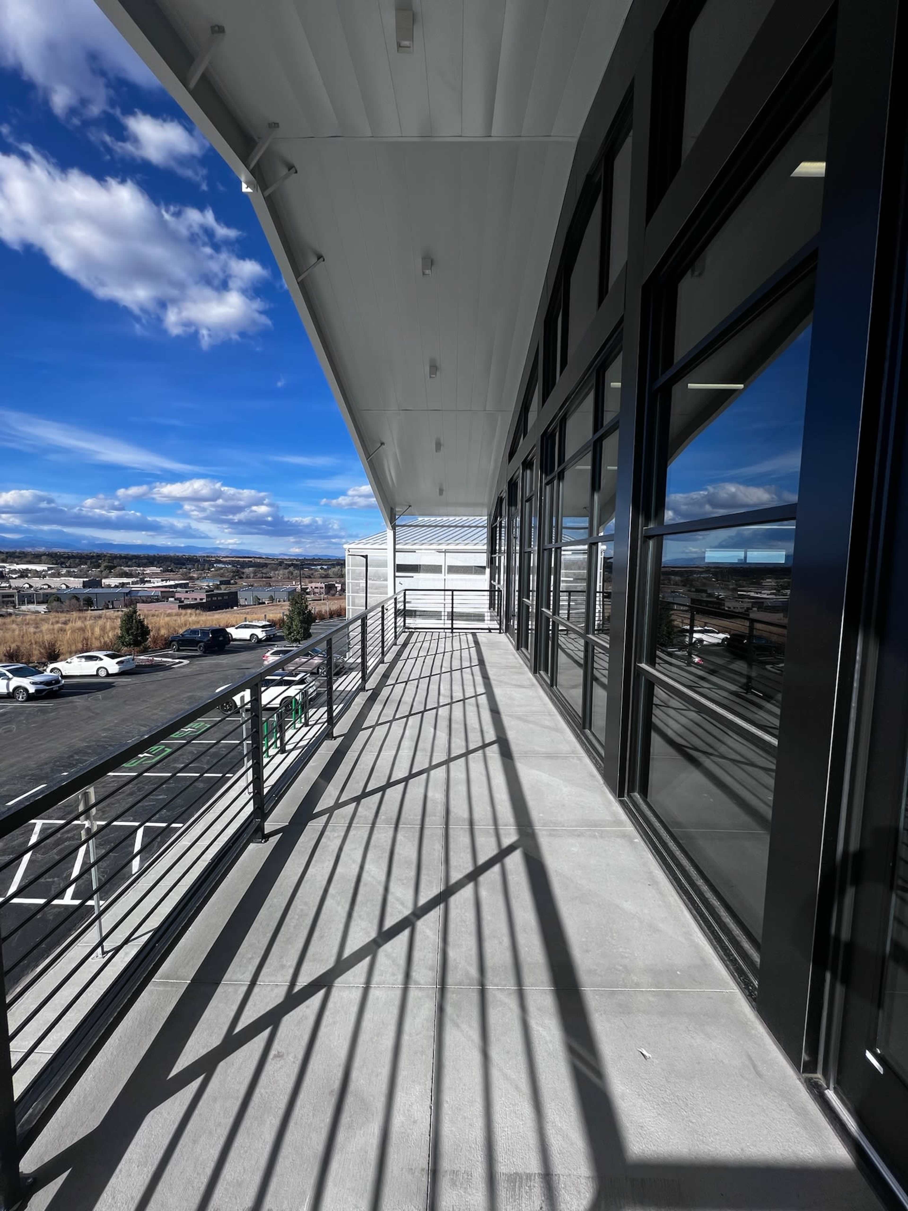 A walkway with a railing extends alongside a building, featuring large windows and a clear blue sky above.