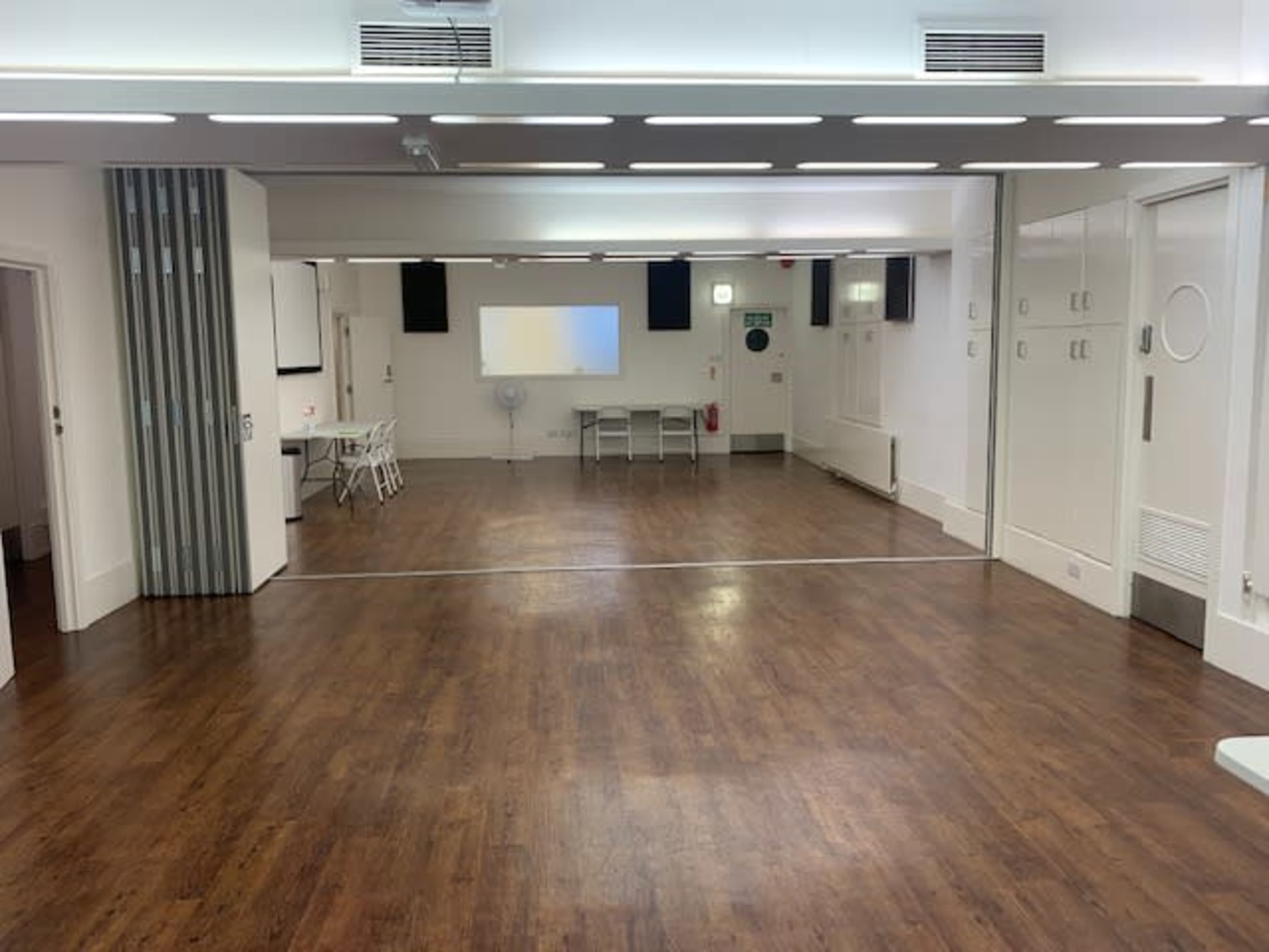 A spacious, empty room with wooden floors, a projector screen on the wall, and a folding table set up in the center.