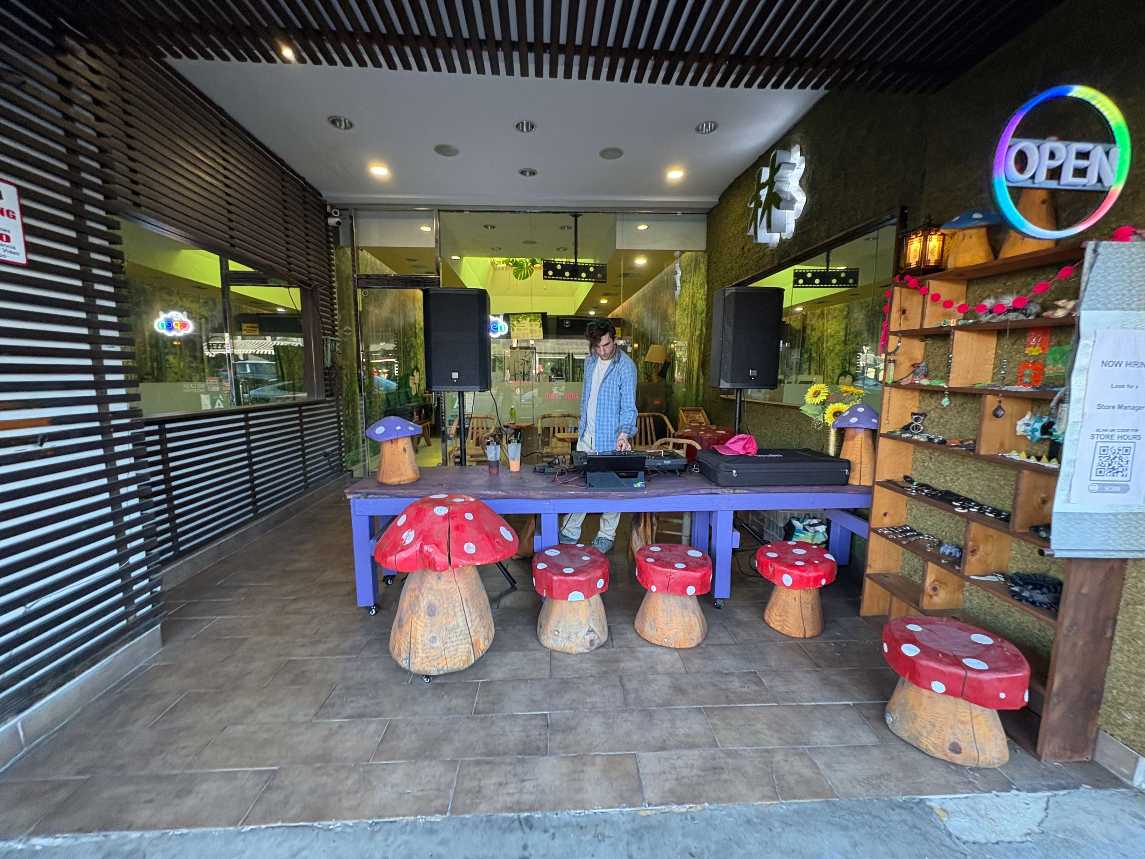 A person stands behind a table in front of a store, surrounded by wooden mushroom-shaped stools and decorative shelves.