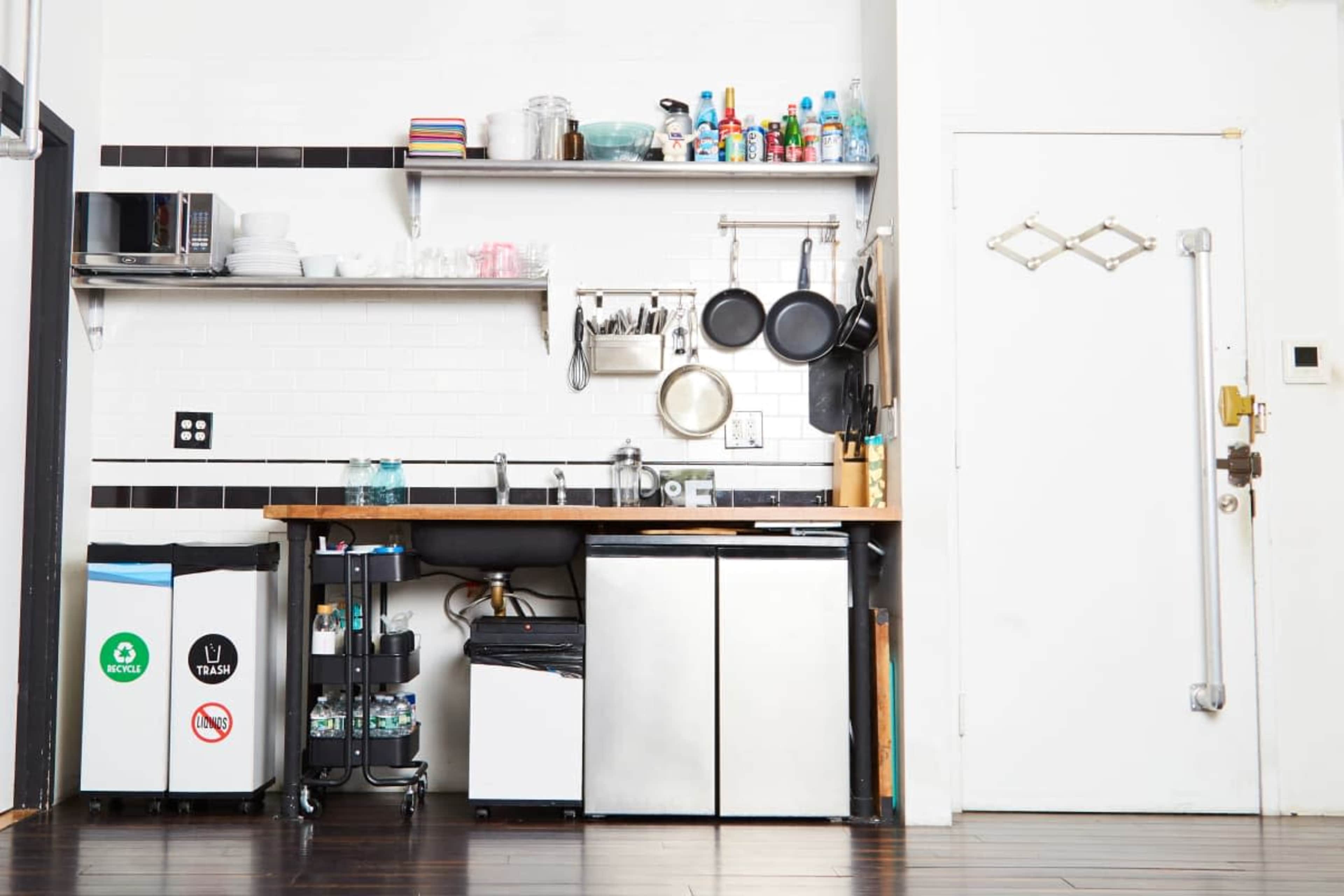 The image shows a neat kitchen area featuring a silver refrigerator, a sink, shelves with various containers, and a door on the right.