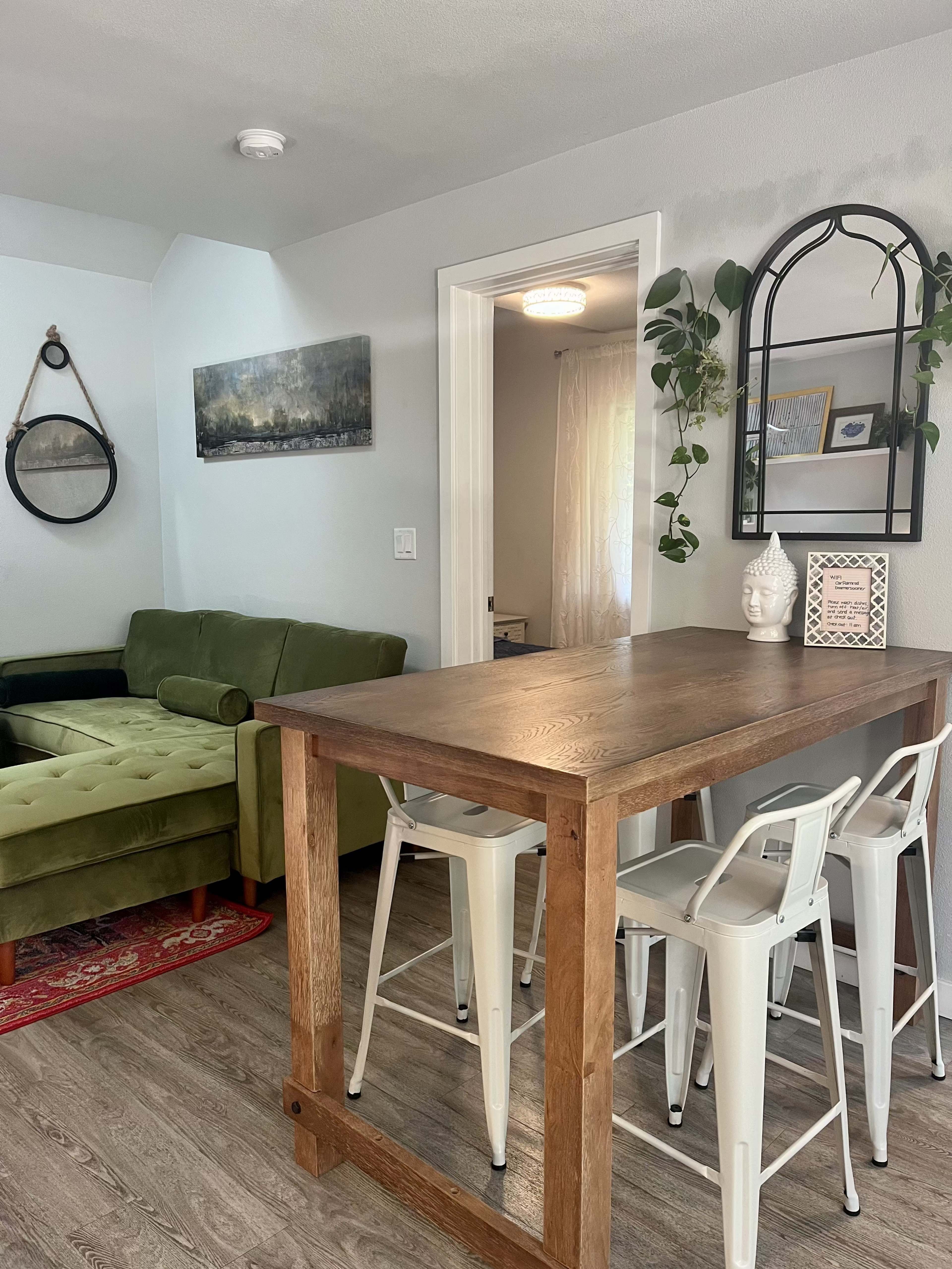 A wooden table with white bar stools is set against a wall in a living area featuring a green couch and a decorative mirror.