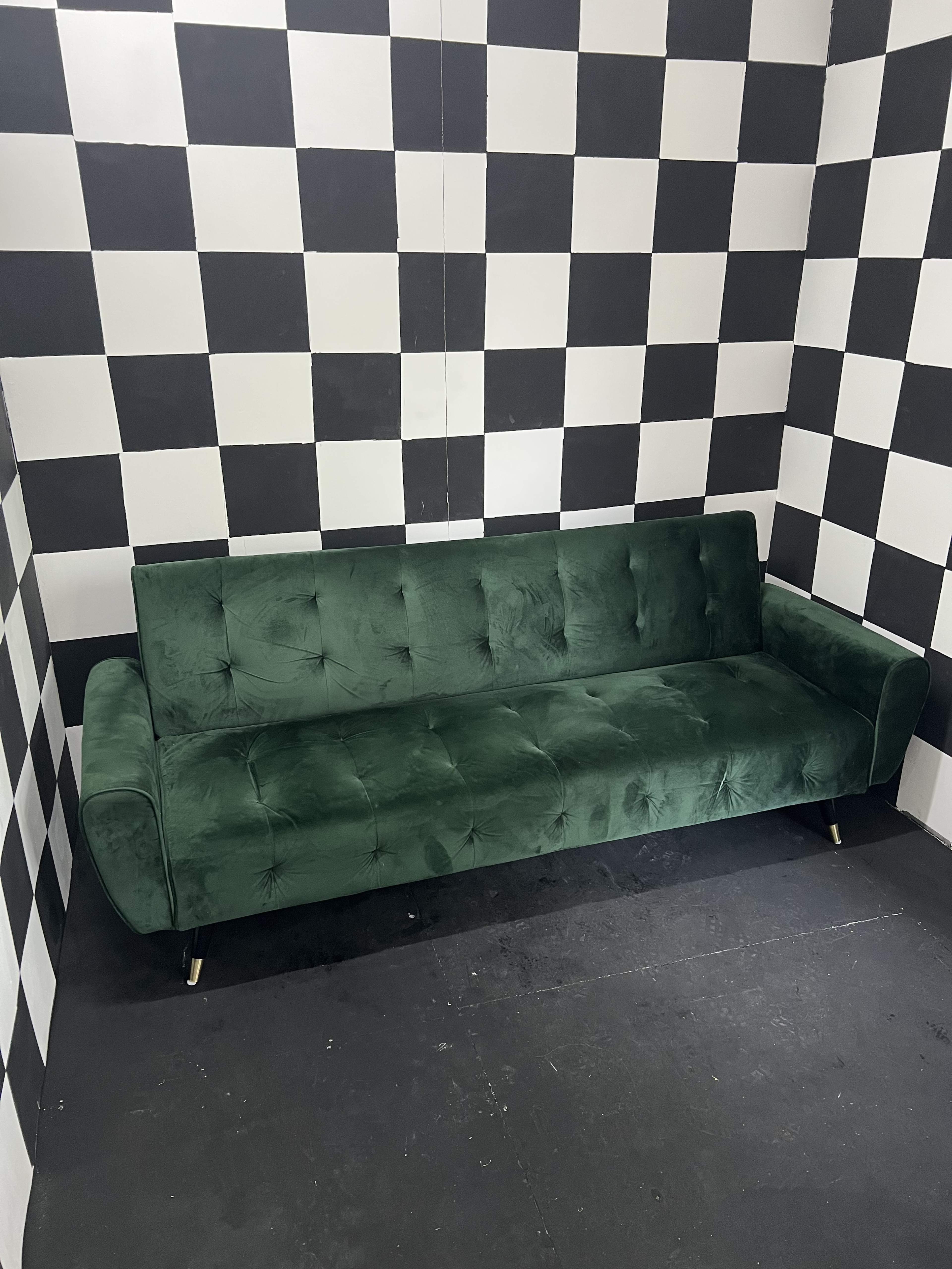 The image shows a green velvet sofa positioned against a black and white checkered wall.