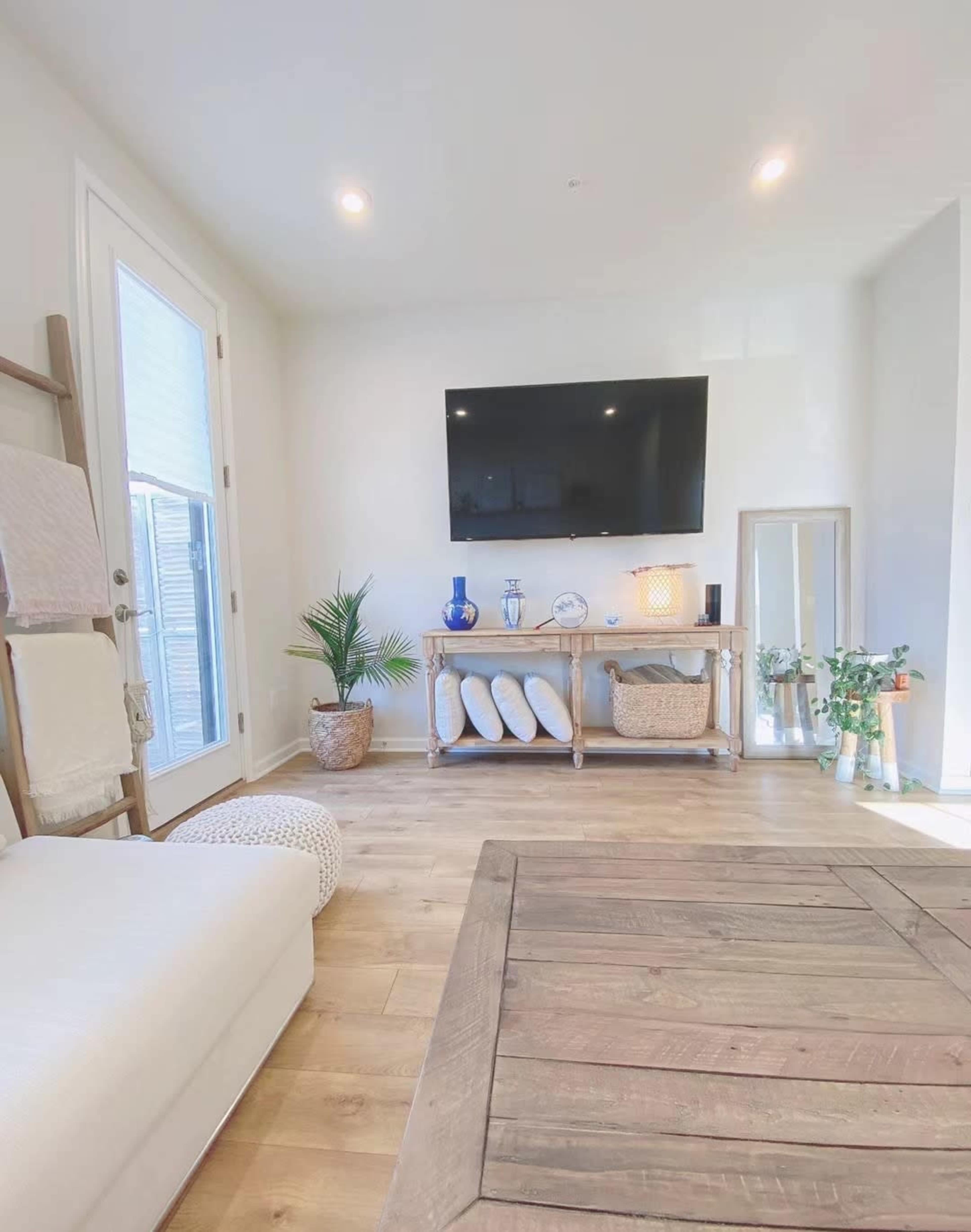 A modern living room features a large television mounted on the wall opposite a wooden console table, with a decorative plant and a mirror leaning beside it.