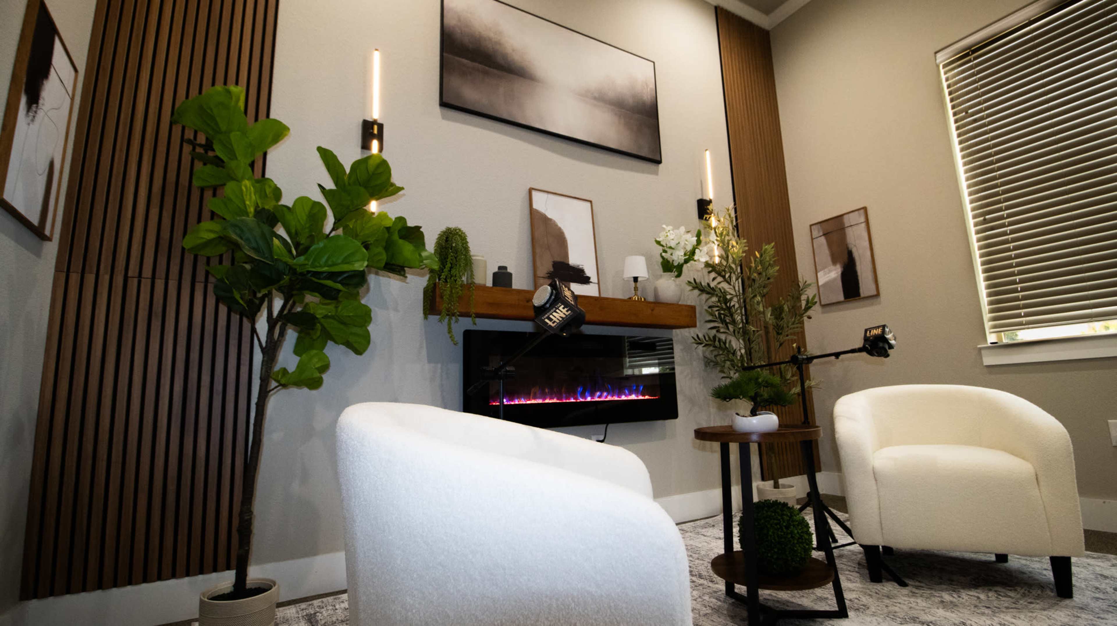 The image shows a cozy indoor space featuring two white armchairs, a modern electric fireplace with a decorative mantel, and potted plants against a backdrop of wooden paneling and framed artwork.