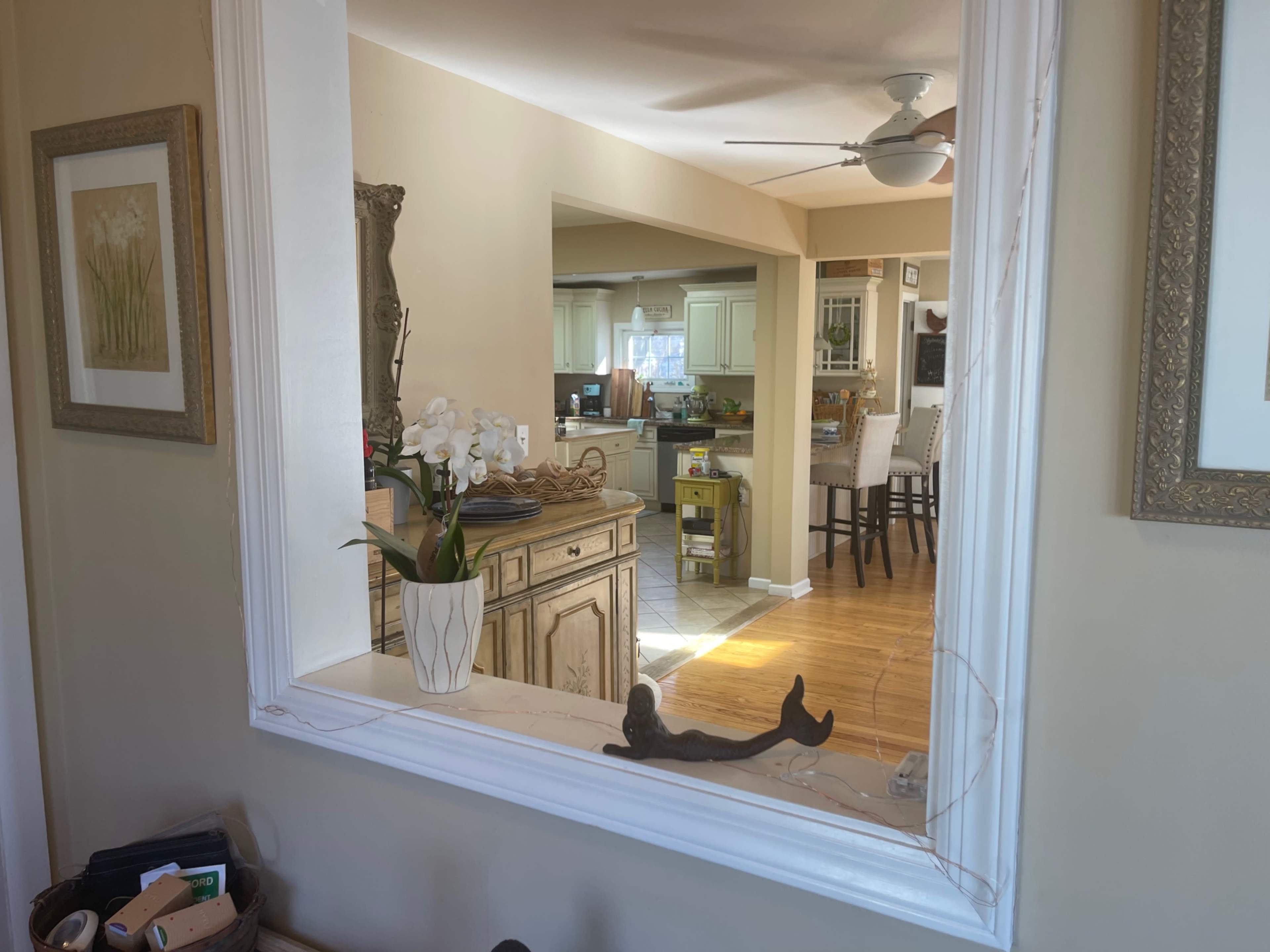 A large decorative mirror frame reflects a view of a kitchen area with a dining table and chairs in the background.
