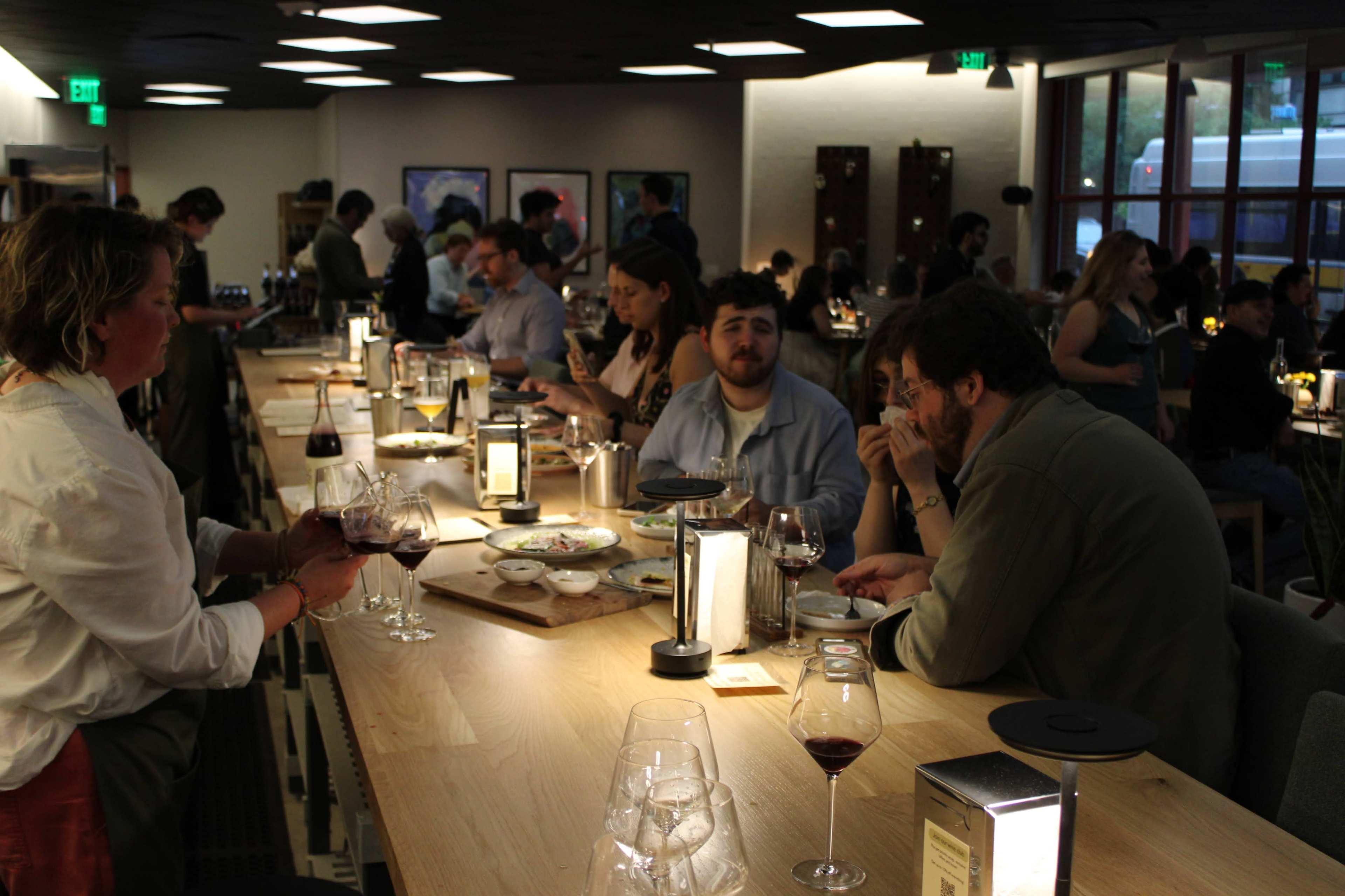 A busy dining area with several guests seated at a long wooden bar, engaged in conversation and enjoying drinks, while a server delivers wine to a table.