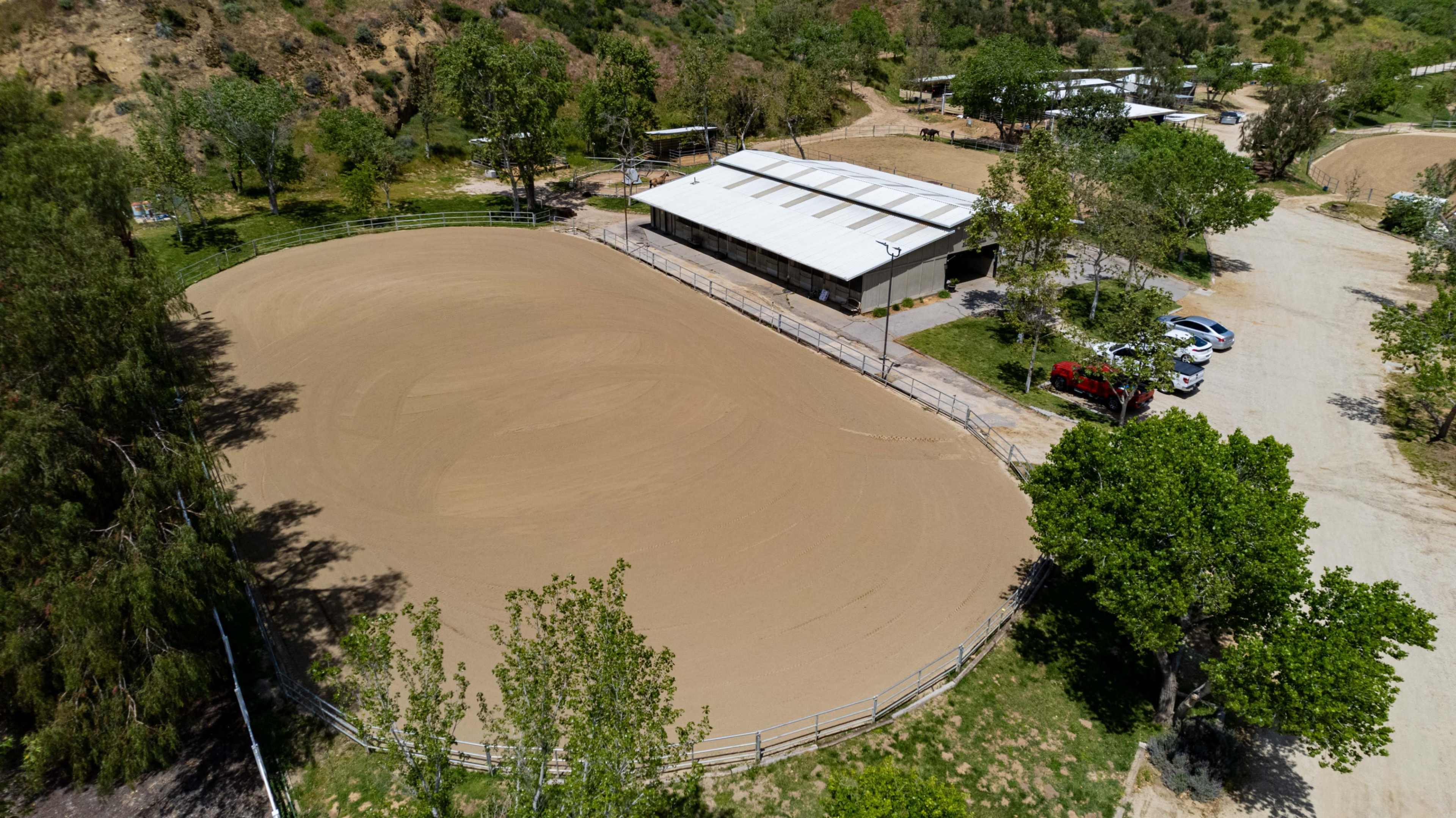 The image shows an aerial view of a large, empty riding arena surrounded by trees and a building, with parked cars nearby.