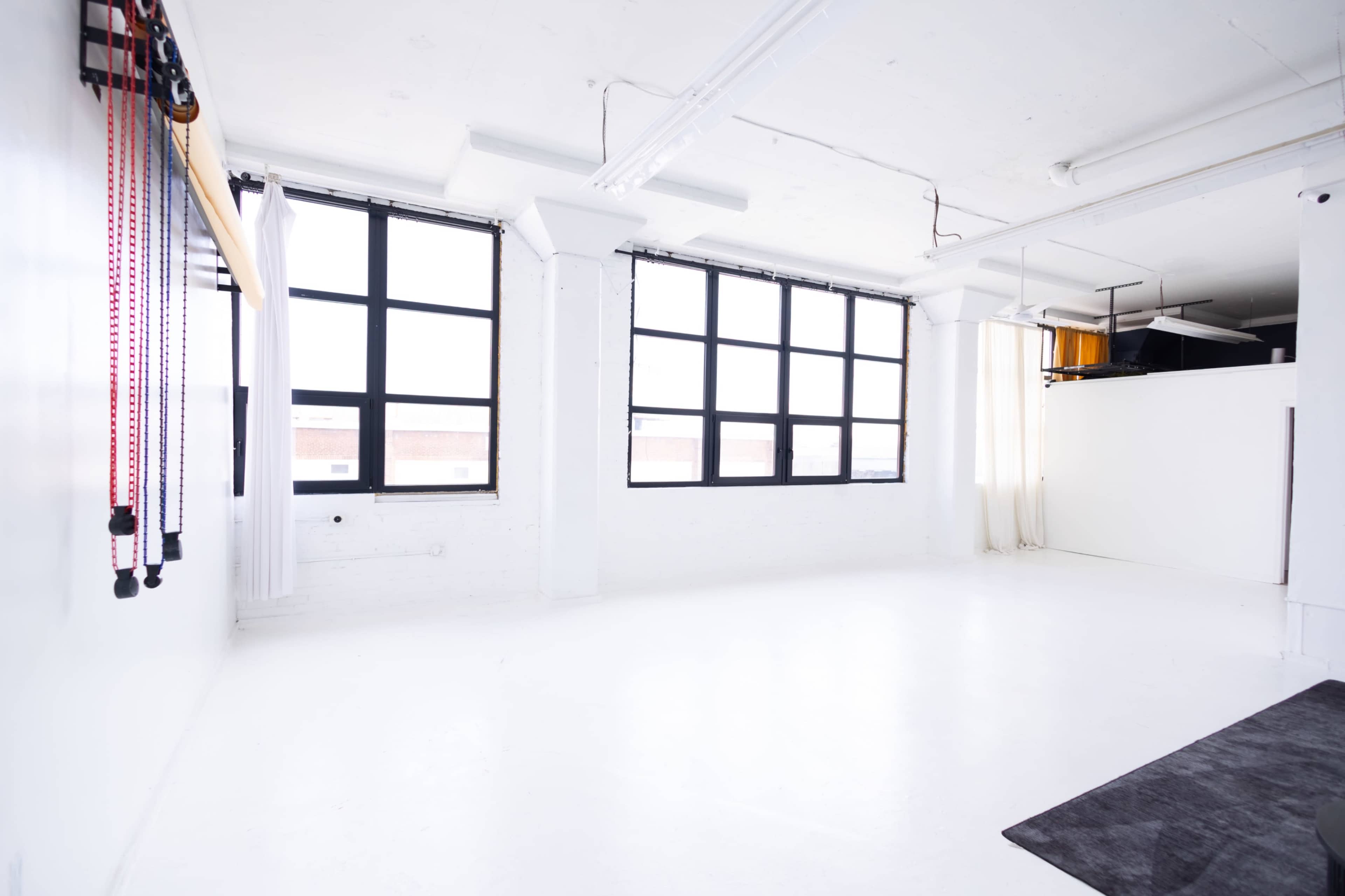 The image shows a spacious, brightly lit room with large windows, white walls, and a minimalistic design.
