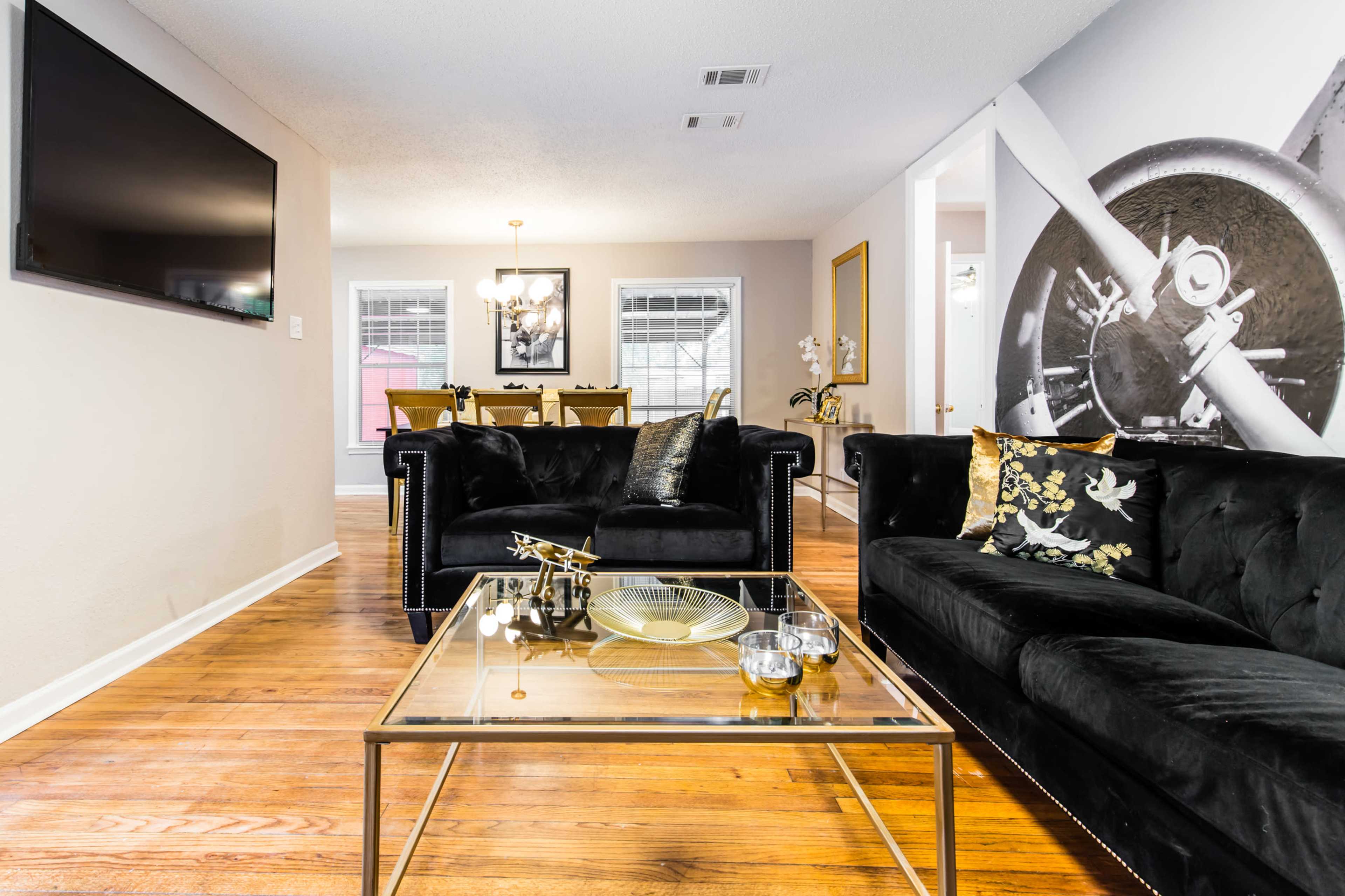 A modern living room with a black velvet couch facing a glass coffee table, accented by a large wall mural of an airplane propeller and a dining area in the background.