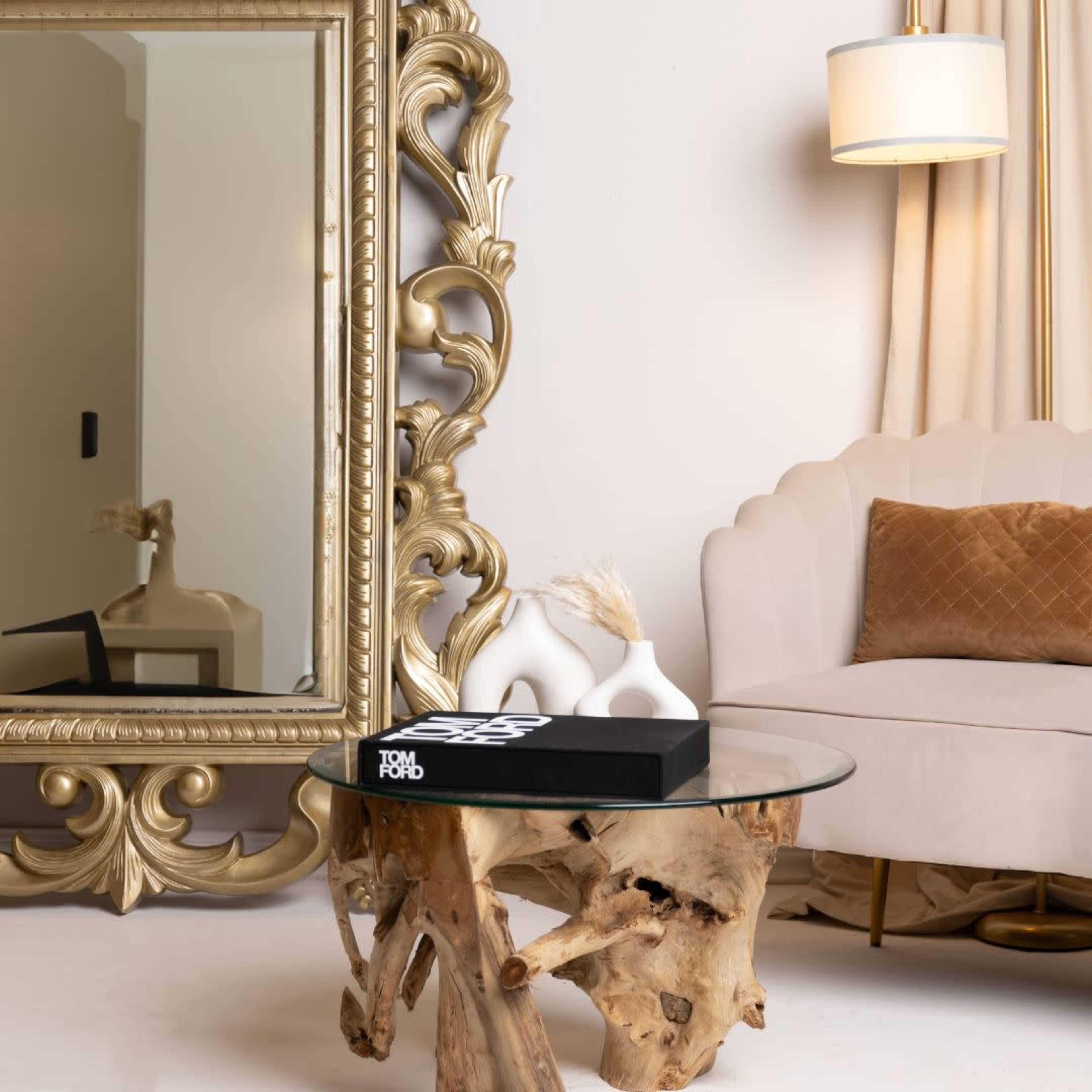 A stylish living room setting with a plush beige sofa next to a round glass table supported by a wooden base, and a large ornate gold-framed mirror on the wall.
