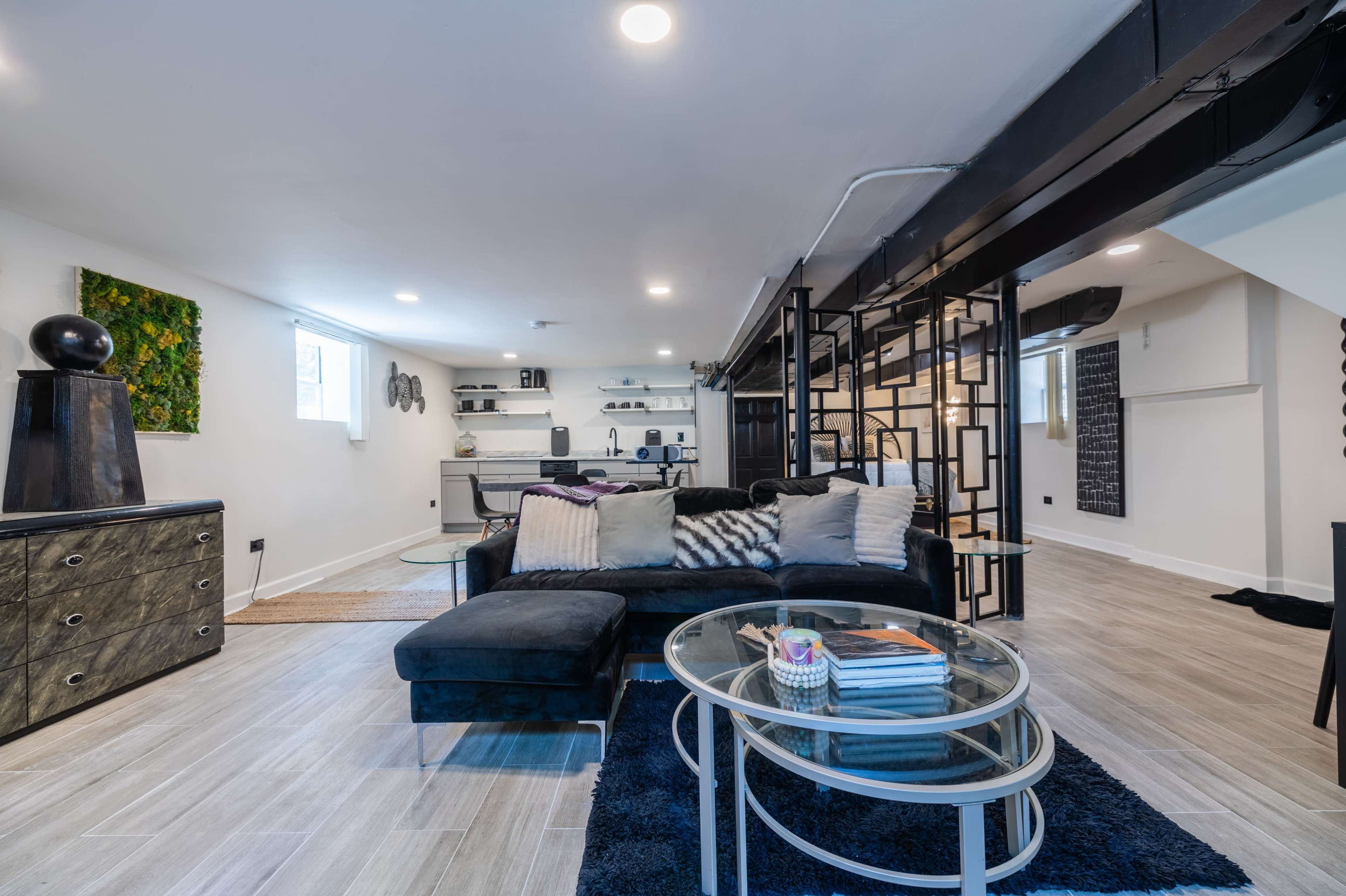 A modern basement living area features a sectional sofa, a glass coffee table, and a workspace, with white walls and wooden flooring.