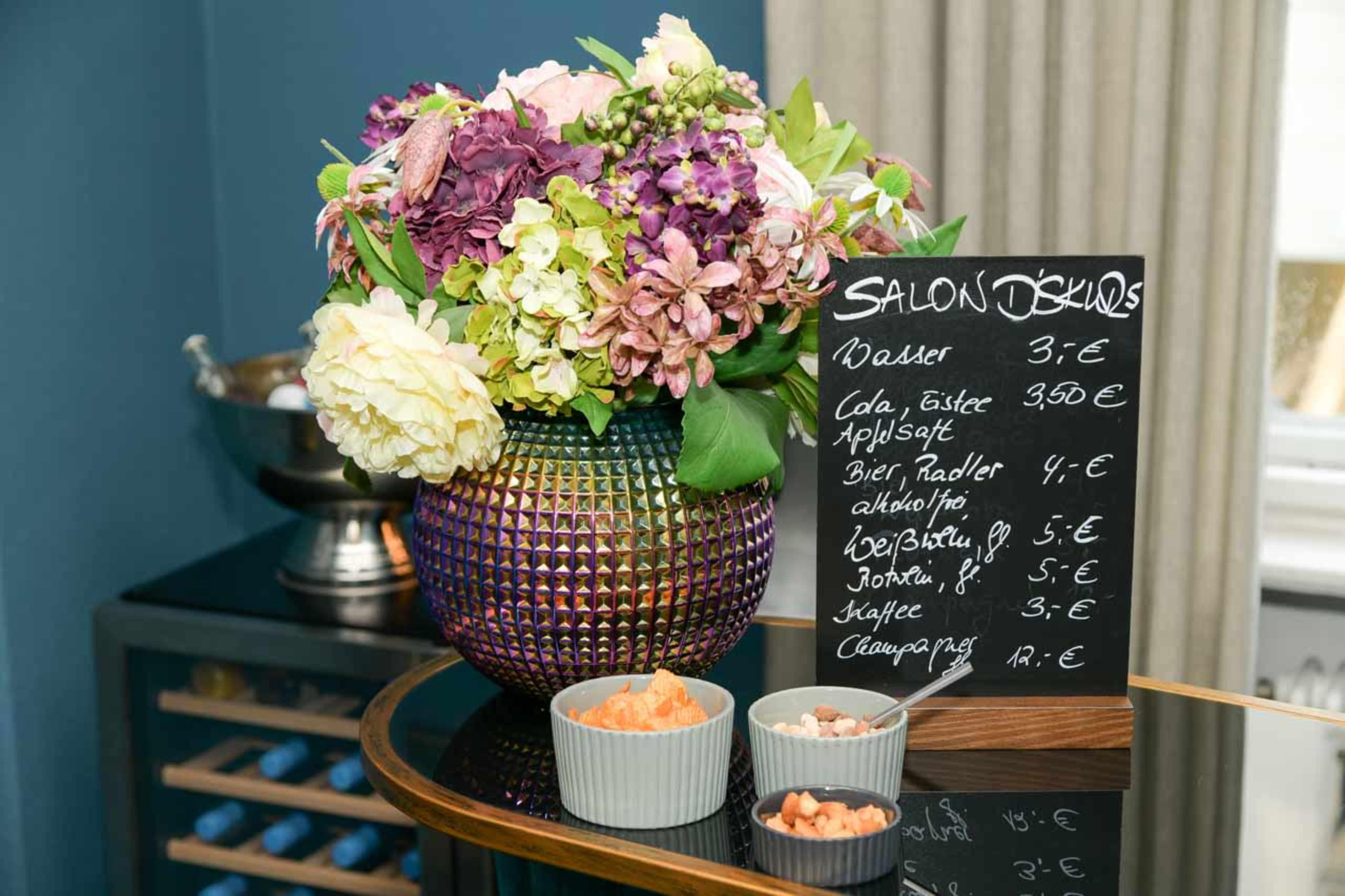 A colorful arrangement of flowers sits next to a chalkboard menu displaying beverage prices and a selection of snacks.