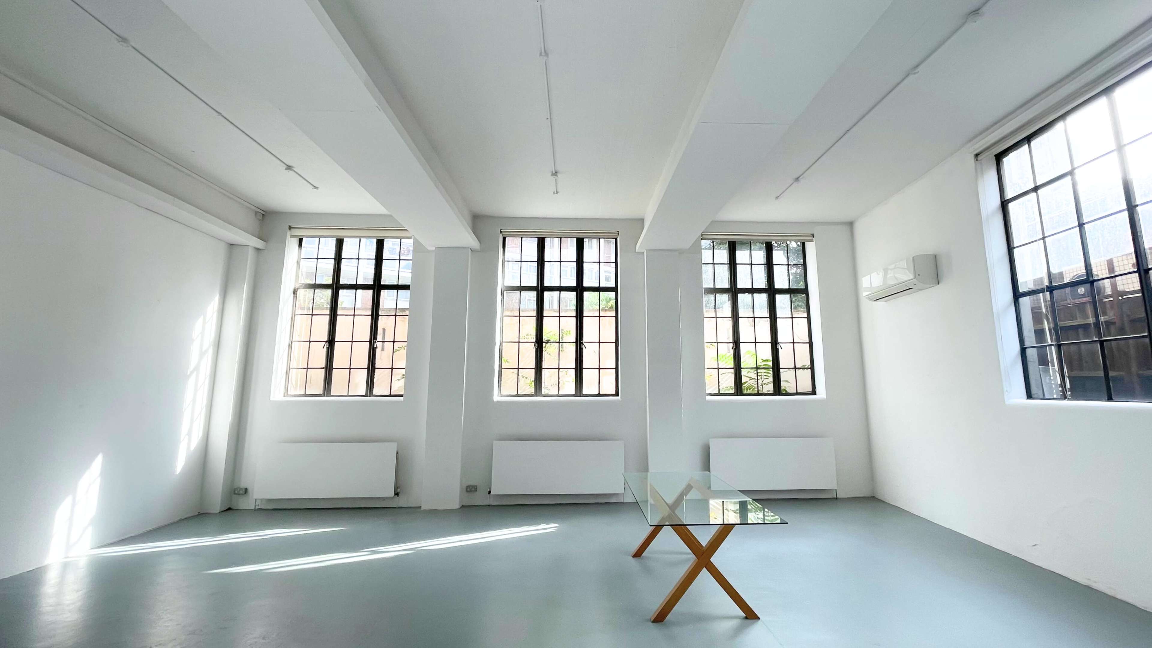 A bright, minimalistic room features three large windows and a glass table with wooden legs in the center.