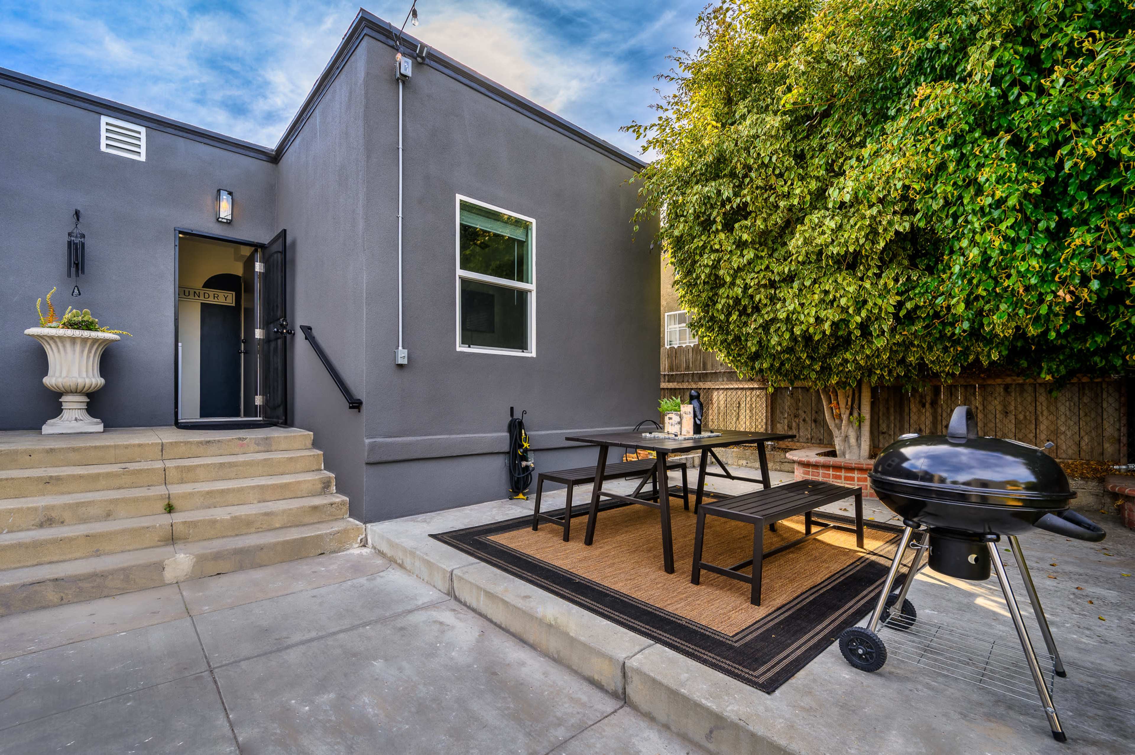 A gray-walled house with a staircase leads to a patio featuring a table and chairs, alongside a charcoal grill and a large bush.