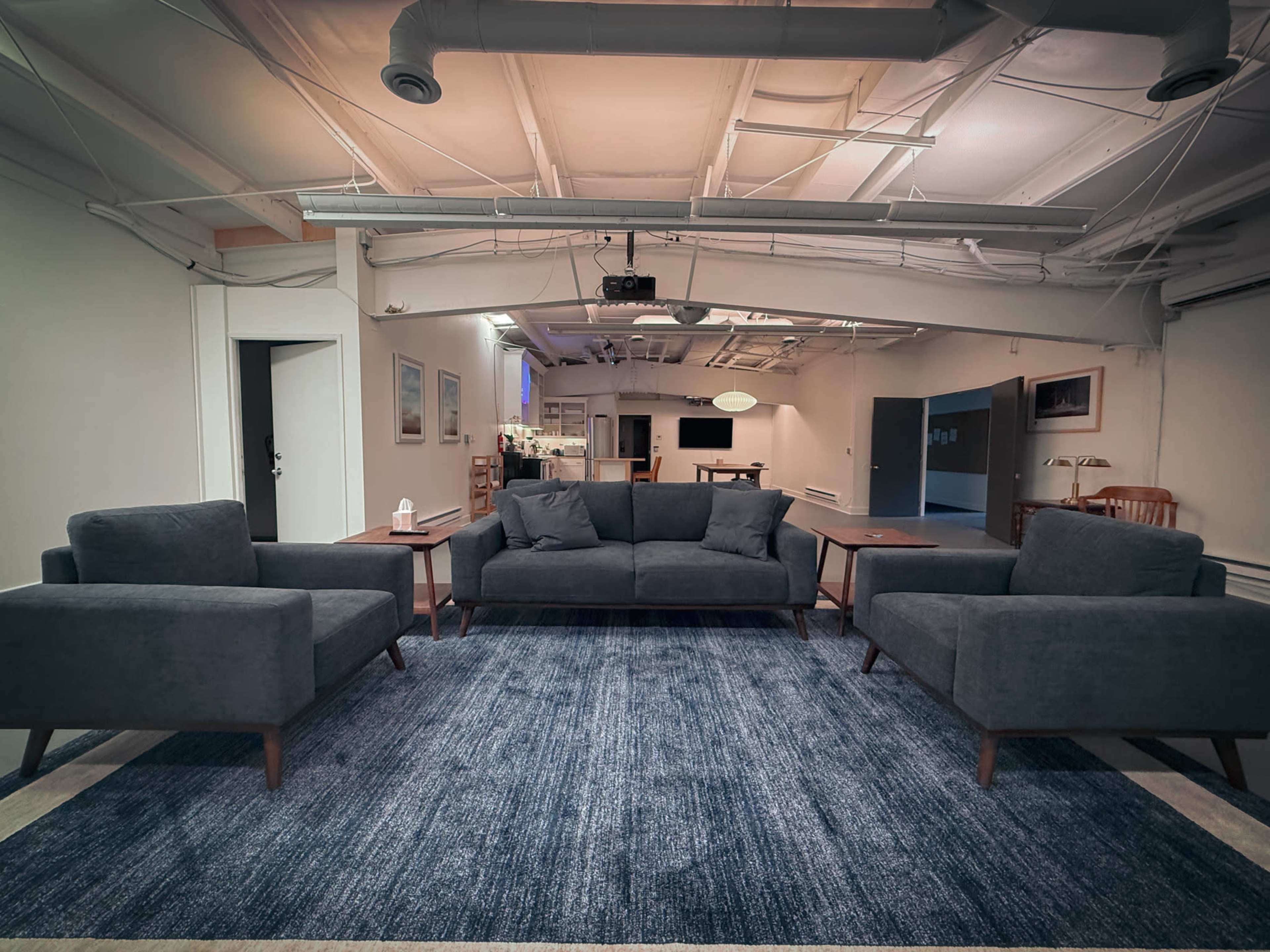 A modern living area features a central dark gray sofa flanked by two matching armchairs, all set on a blue rug with a spacious, minimalist backdrop.