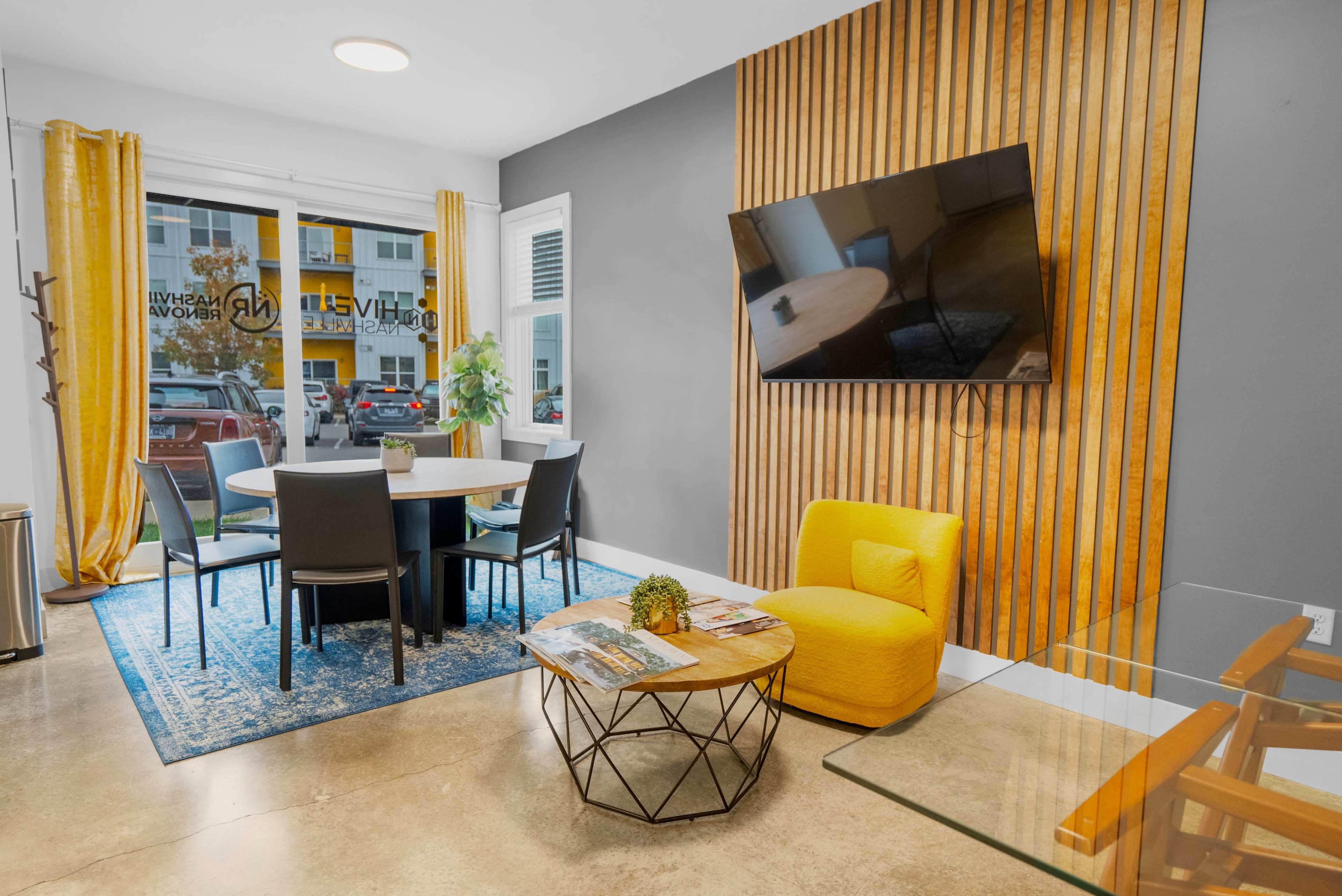 A modern sitting area with a round table and chairs, a yellow accent chair, a wooden wall, and a television mounted on the wall.