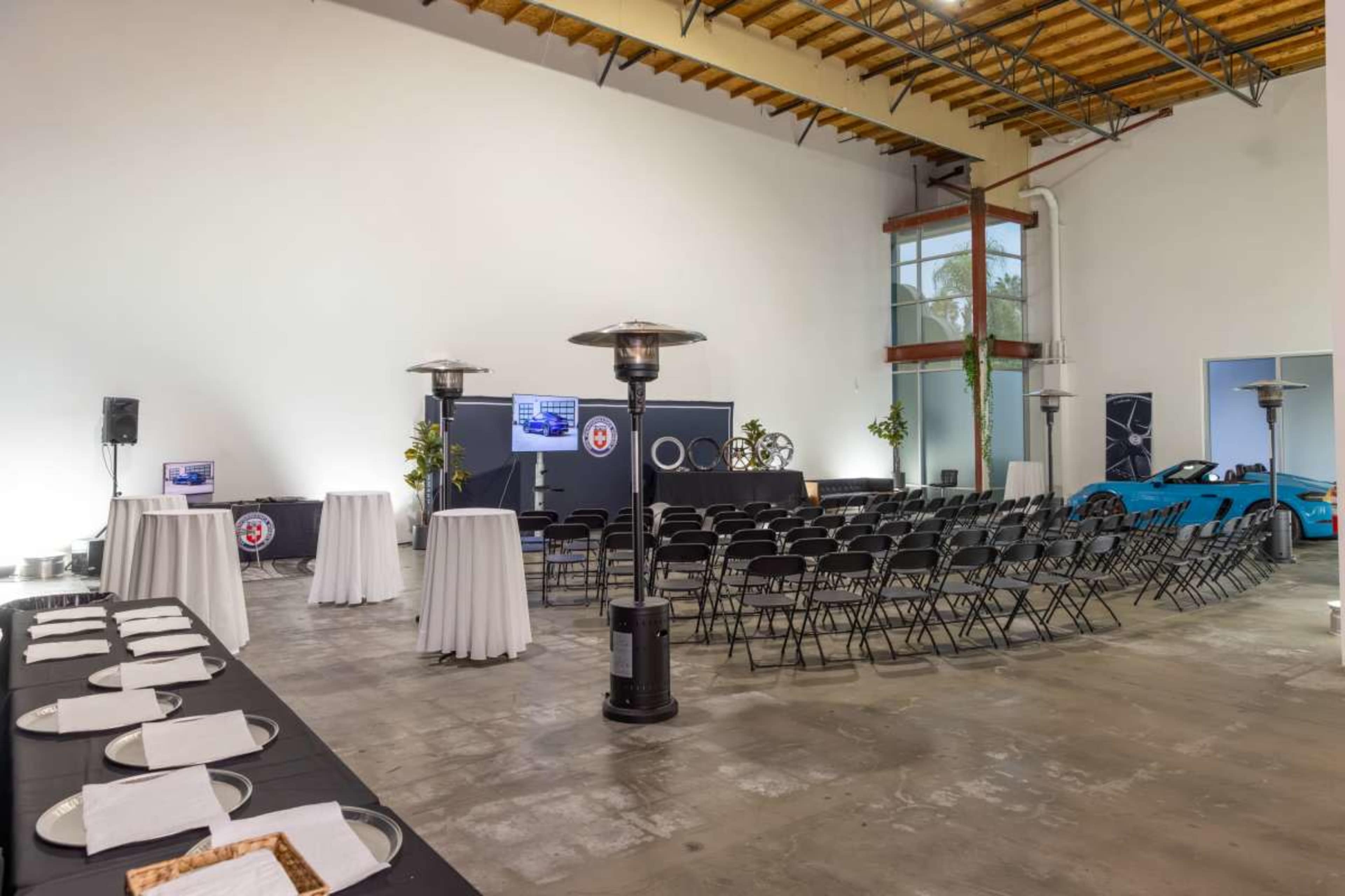 The image shows an event space with rows of black chairs, a table set with white linens, and a blue sports car parked nearby.
