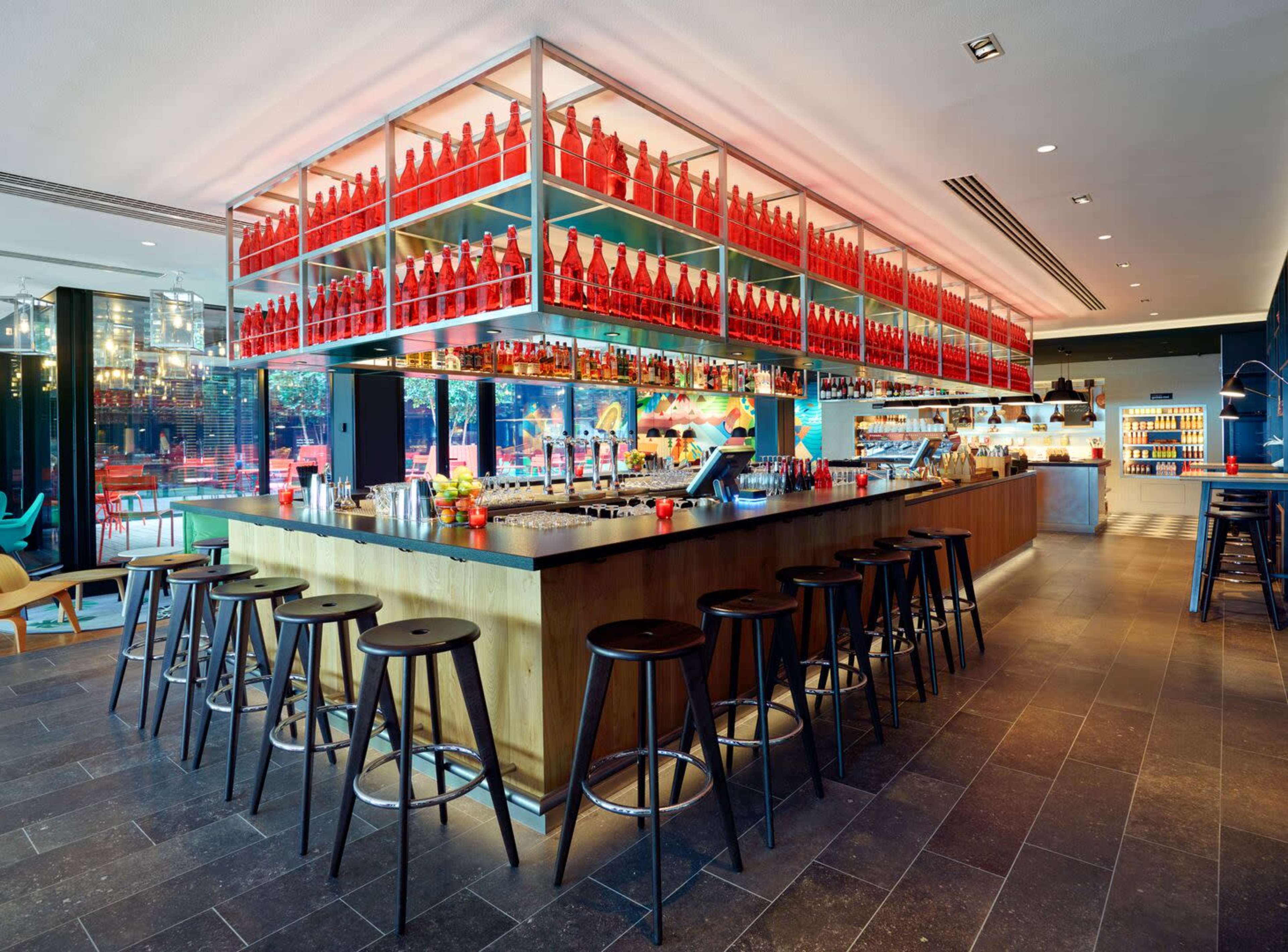 A modern bar features a central wooden counter with black stools and is topped with shelves filled with red bottles, surrounded by glass walls and colorful seating.